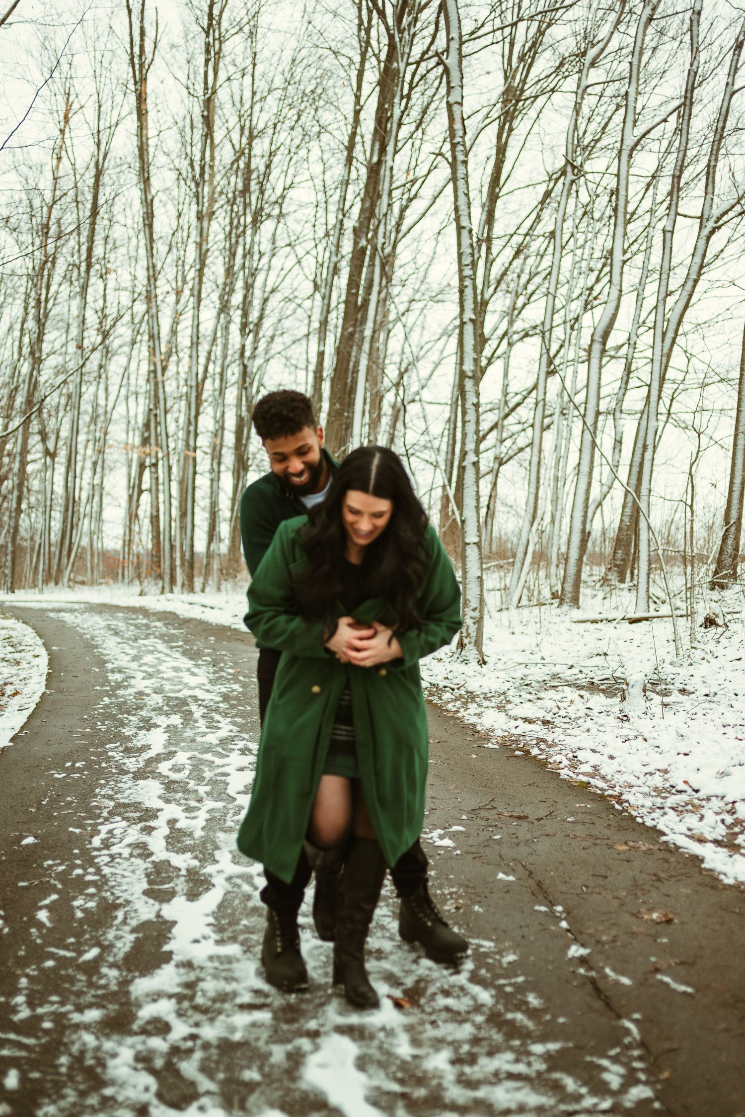 A couple in green winter coats playfully interacts on a snowy forest path in a series of connected photos.