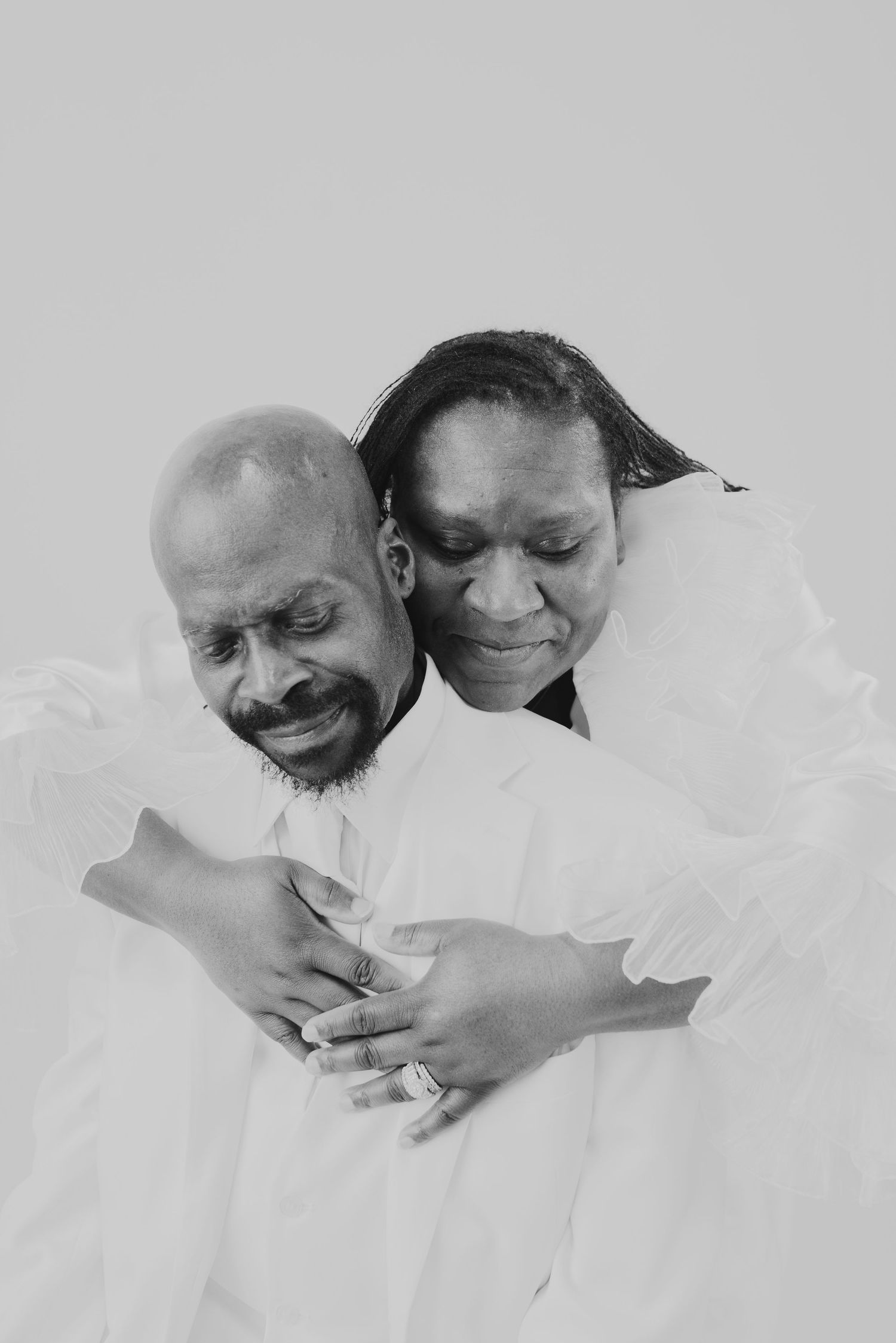 Black and white portrait of a couple embracing tenderly, woman resting her head on mans shoulder, both wearing white.