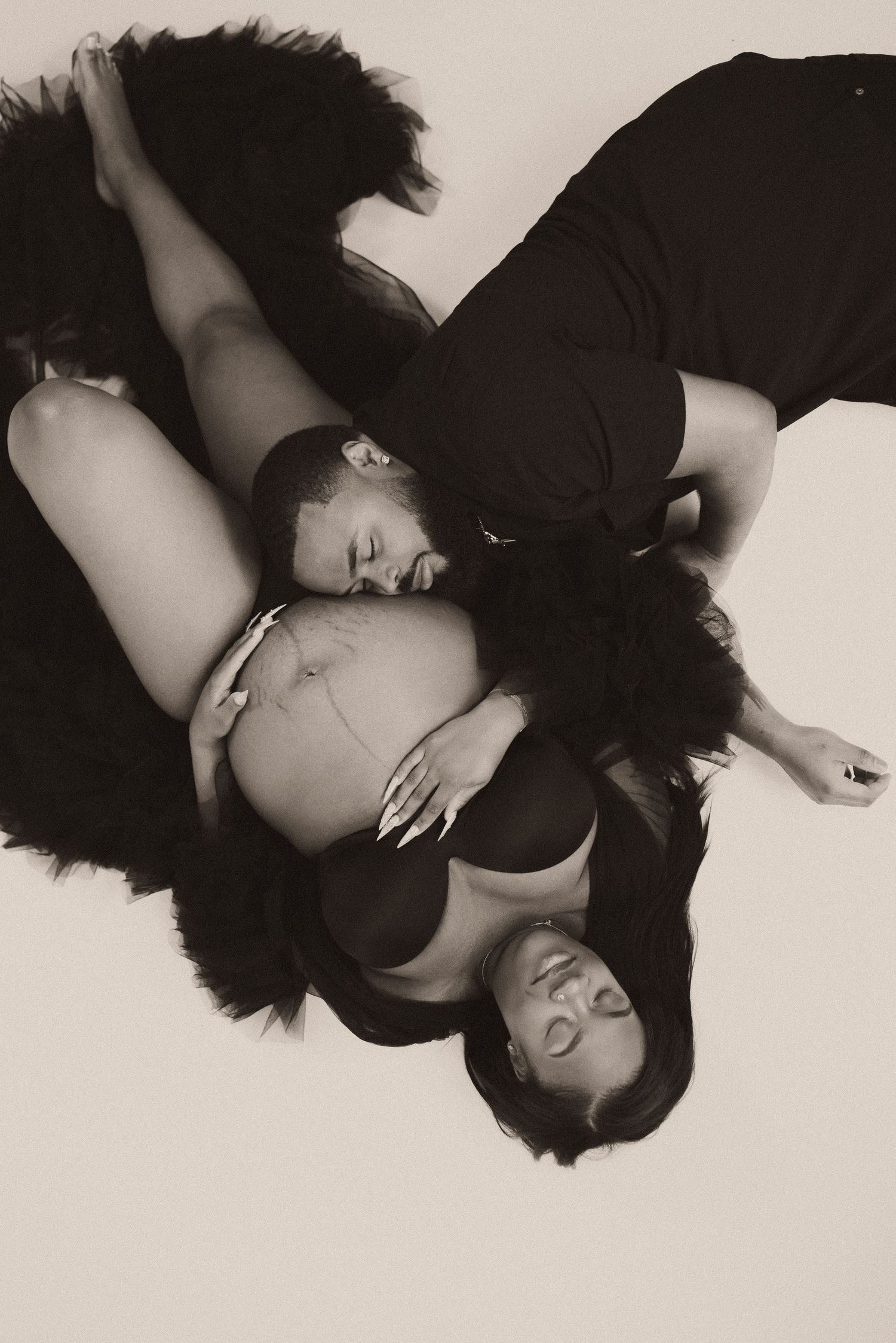 Black and white maternity photo of a couple lying down, man kissing pregnant womans belly, studio portrait.