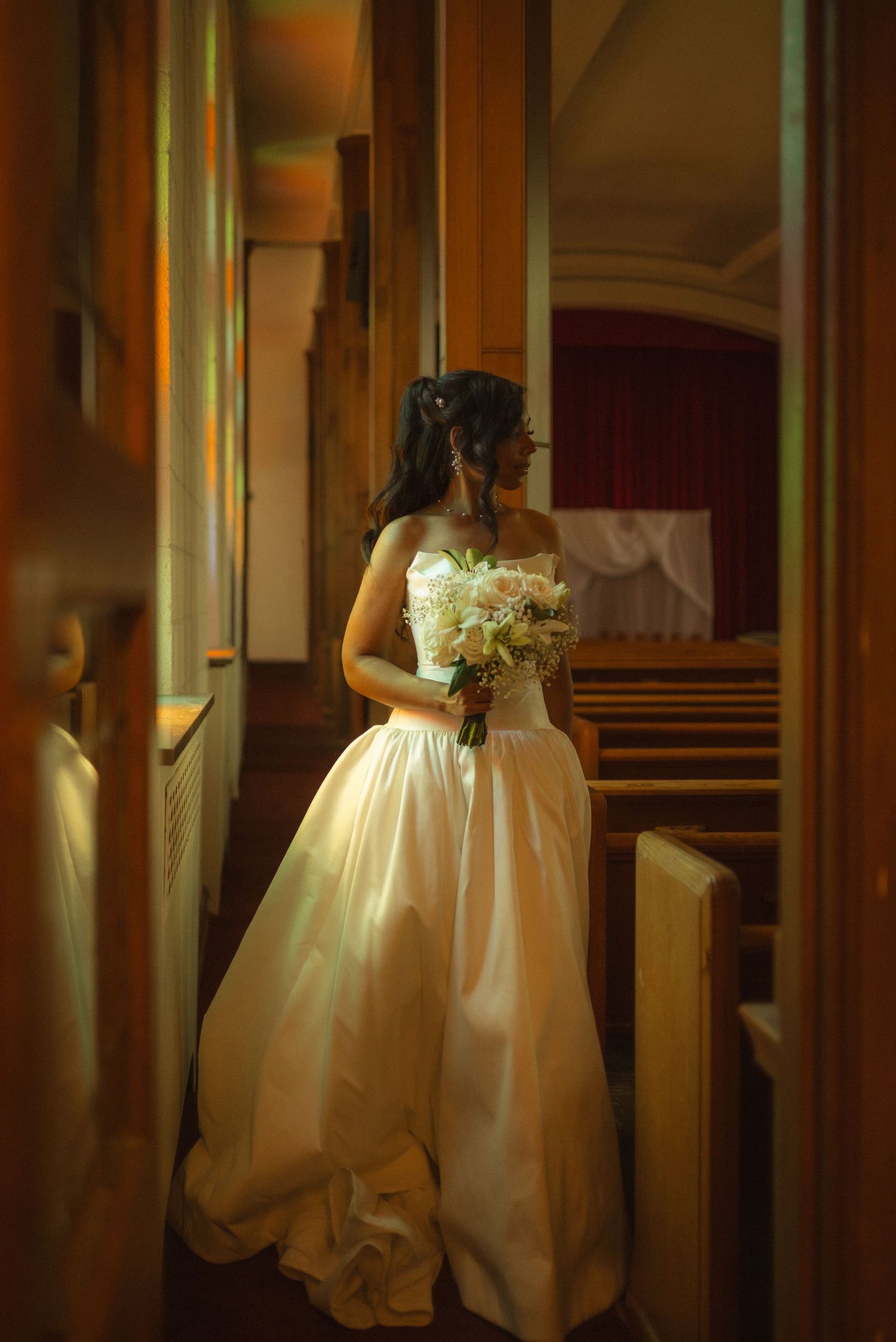 Elegant bride in a strapless ball gown holding a bouquet inside a warmly lit church before her wedding ceremony.