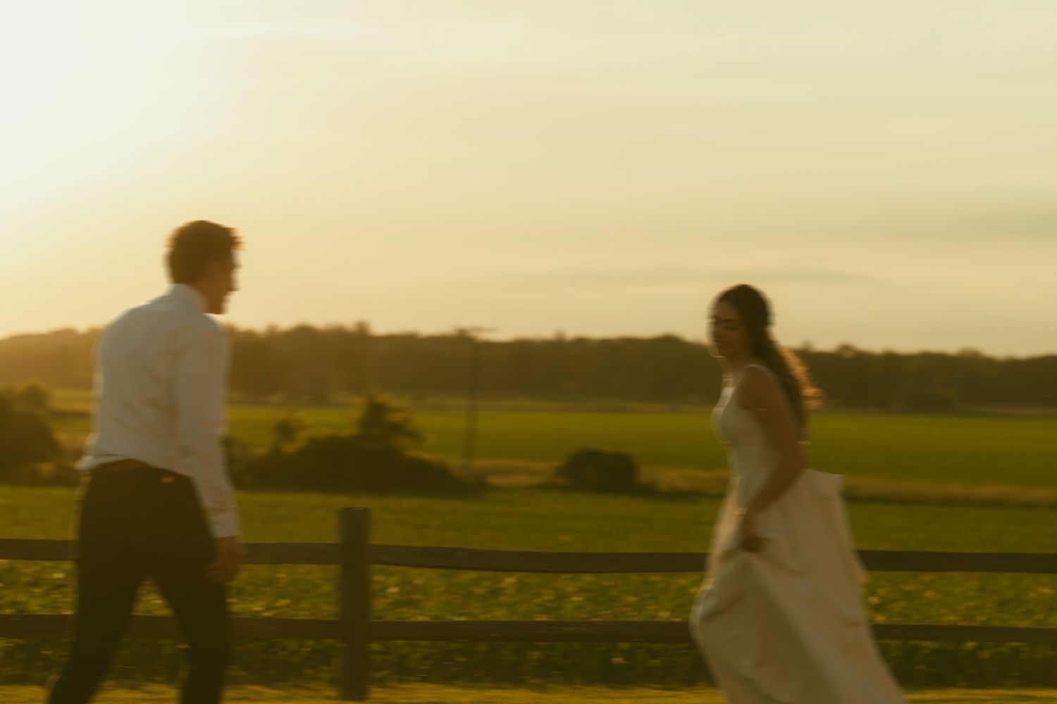 A bride and groom walk through golden sunset light beside a wooden fence with lush green fields in the background.