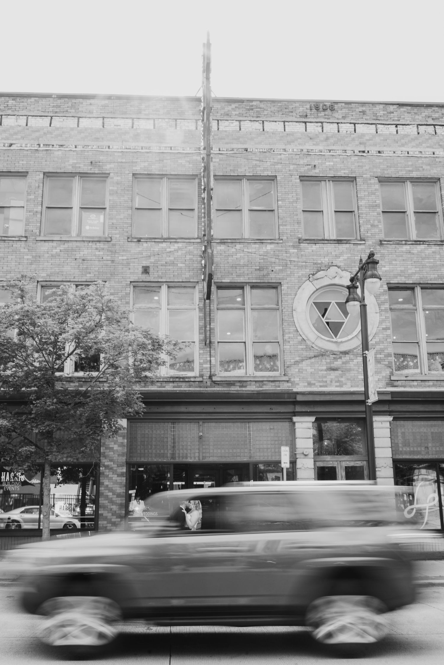 Black and white photo of a blurred car passing a historic brick building with large windows on a city street.