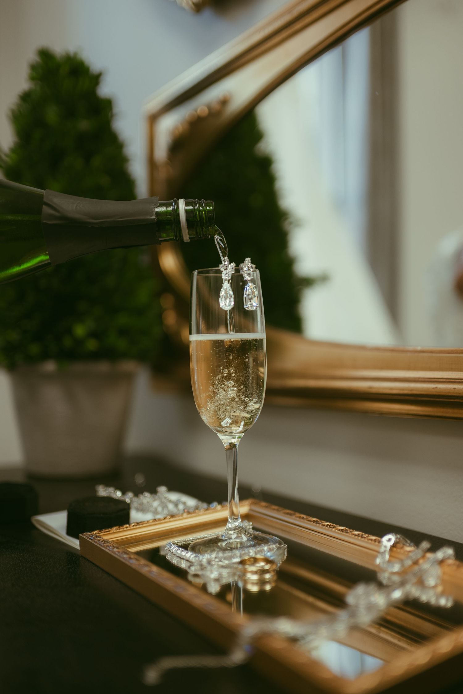 Champagne being poured into a flute glass adorned with diamond earrings on a jewelry tray near a gold mirror.
