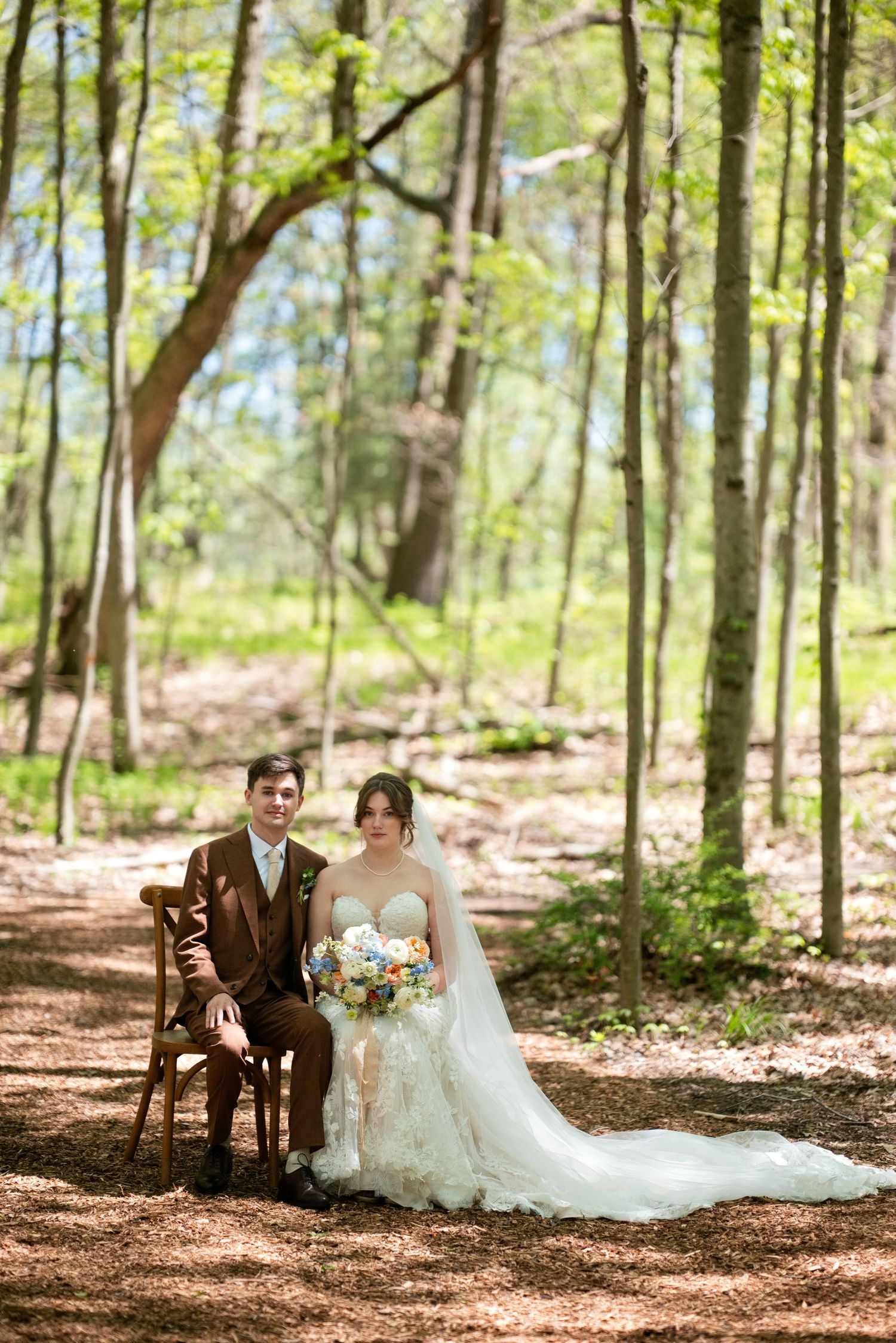 Bride and groom pose in a sunlit forest, she in a white gown with bouquet, he in a brown suit on a wooden chair.