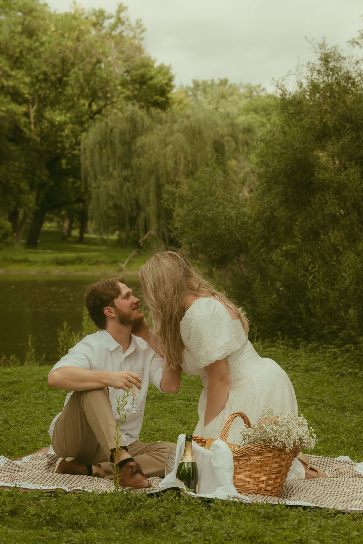 Couple sharing a romantic picnic by a serene lake, surrounded by lush greenery and willow trees.
