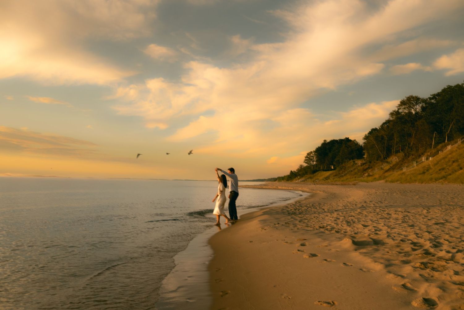 A couple walks along a sandy beach at sunset with dramatic orange clouds in the sky.