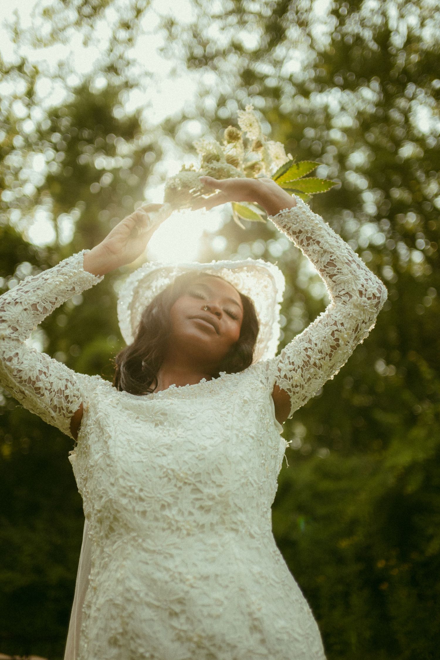 Black bride in white lace dress holds flowers overhead, bathed in golden sunlight among green trees.