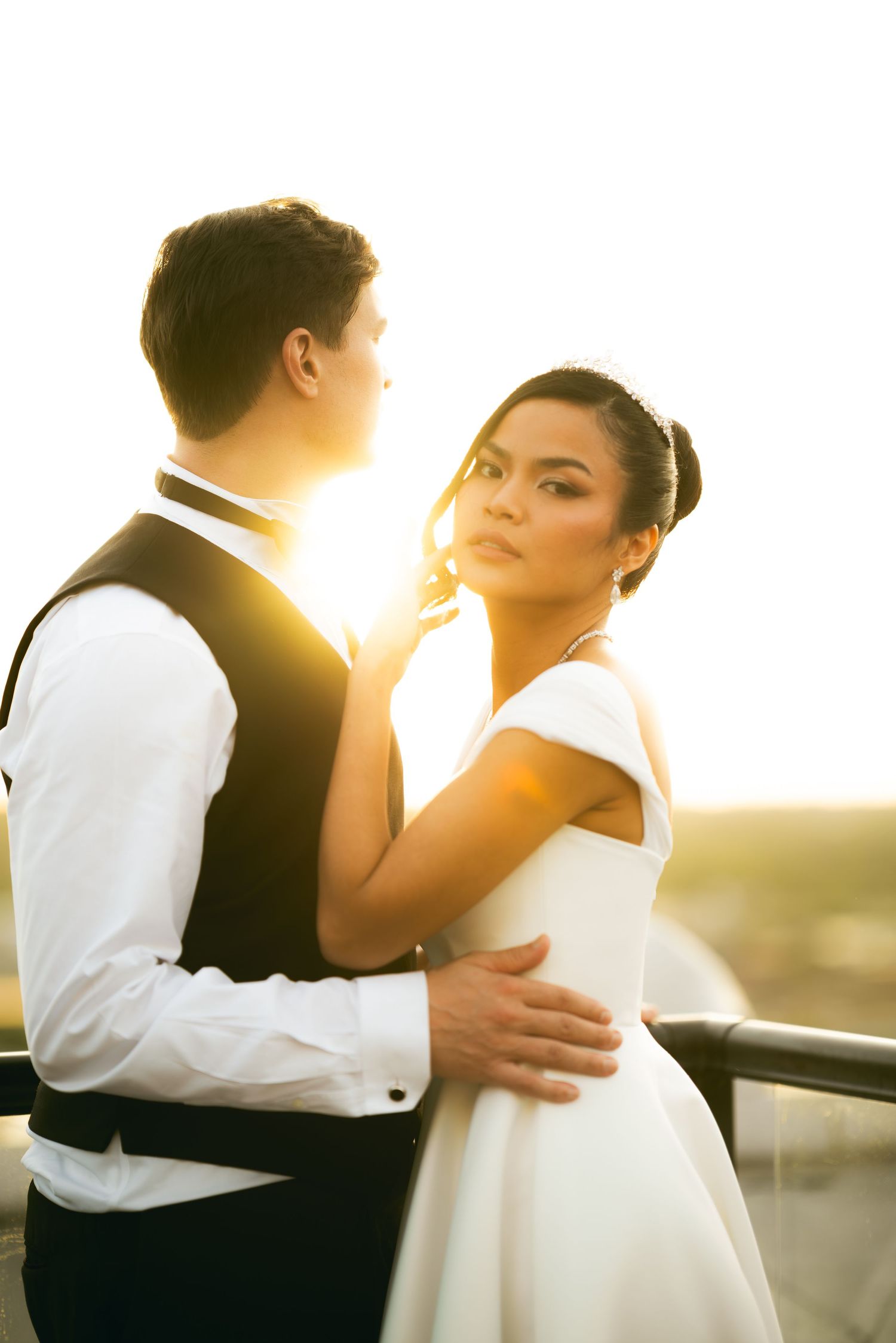 Bride and groom embrace at sunset on a balcony, golden backlight glowing behind them in a romantic wedding portrait.