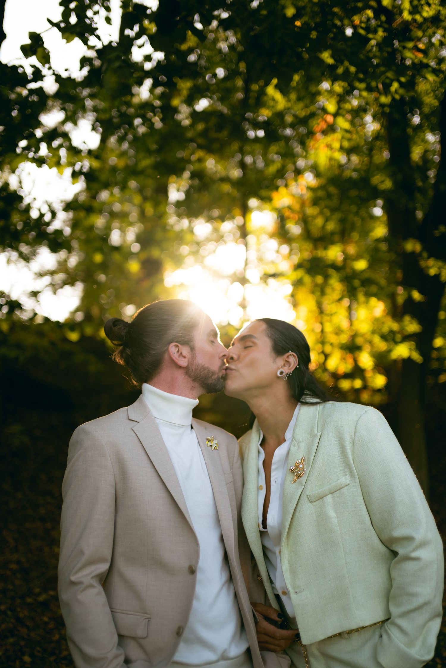 A couple in light colored formal attire share a romantic kiss outdoors with sunlit trees in the background.