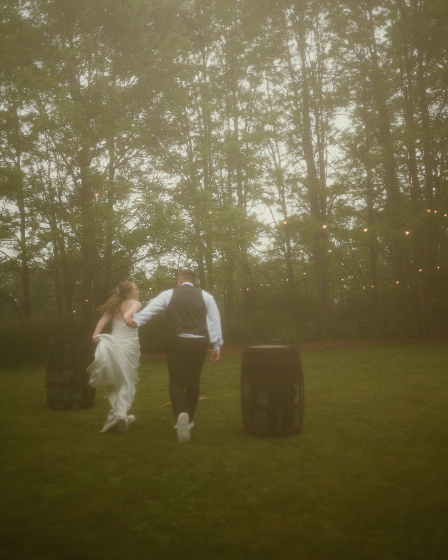 A bride and groom walk hand-in-hand across a misty green lawn surrounded by tall trees and warm string lights at dusk.