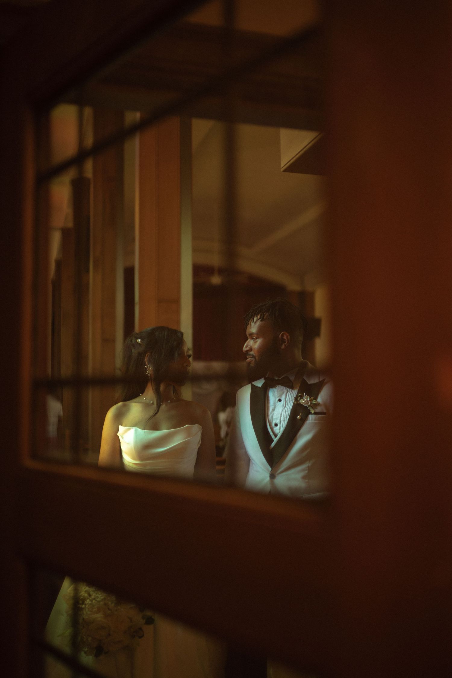 A bride and groom share an intimate moment, viewed through a warmly lit window frame.