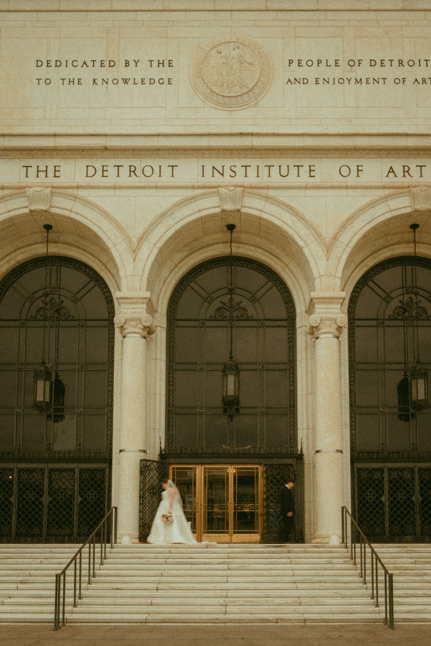 Bride in white gown on steps of Detroit Institute of Arts, elegant neoclassical architecture backdrop.