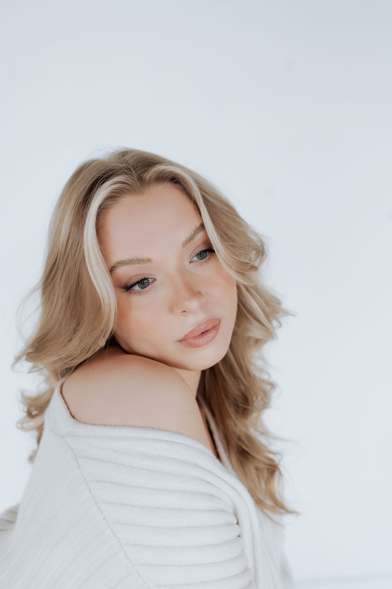 Soft portrait against white background featuring blonde wavy hair and natural makeup in a white knit sweater.