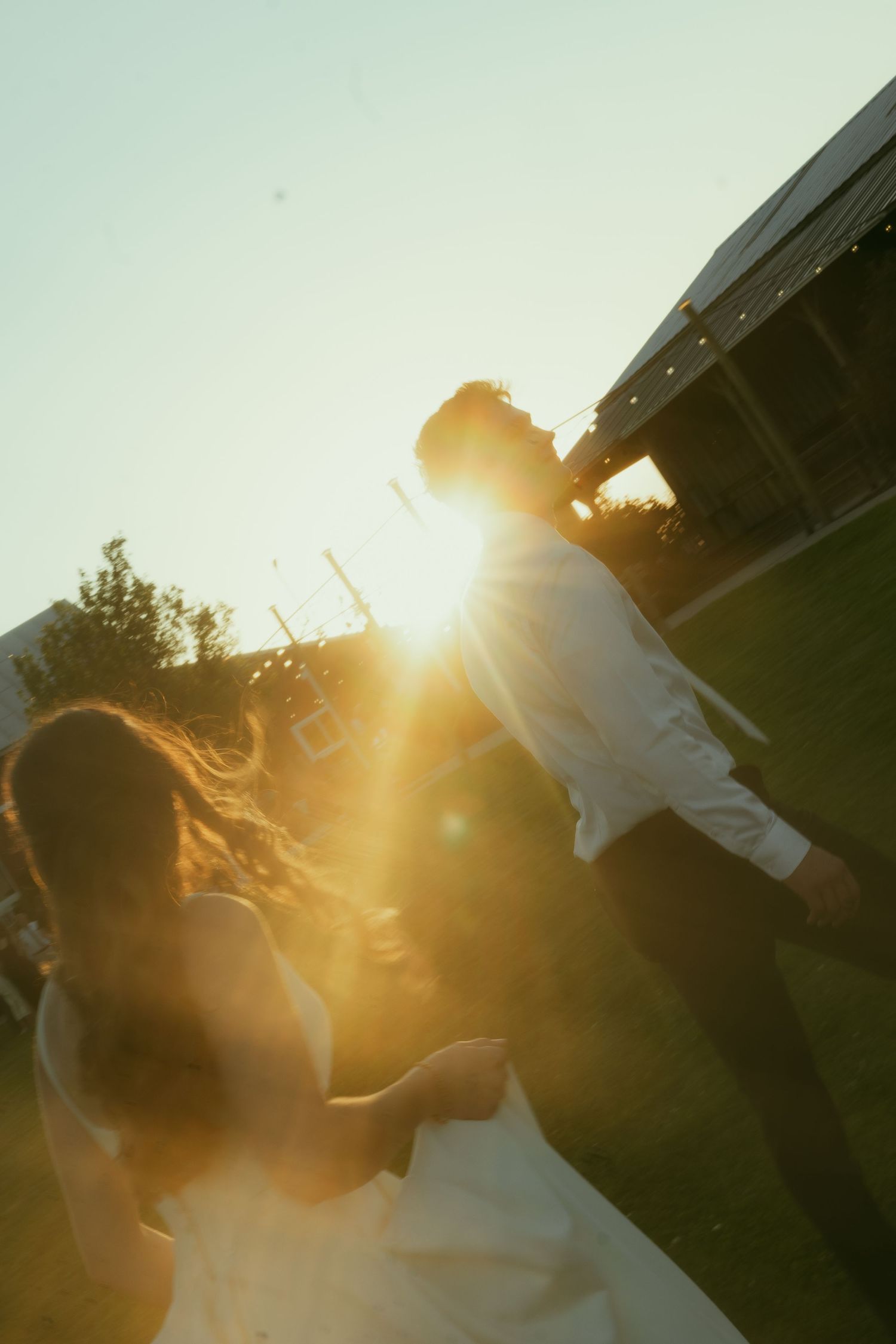 Couple in wedding attire running toward golden sunset light outdoors, backlit with warm sun flare.