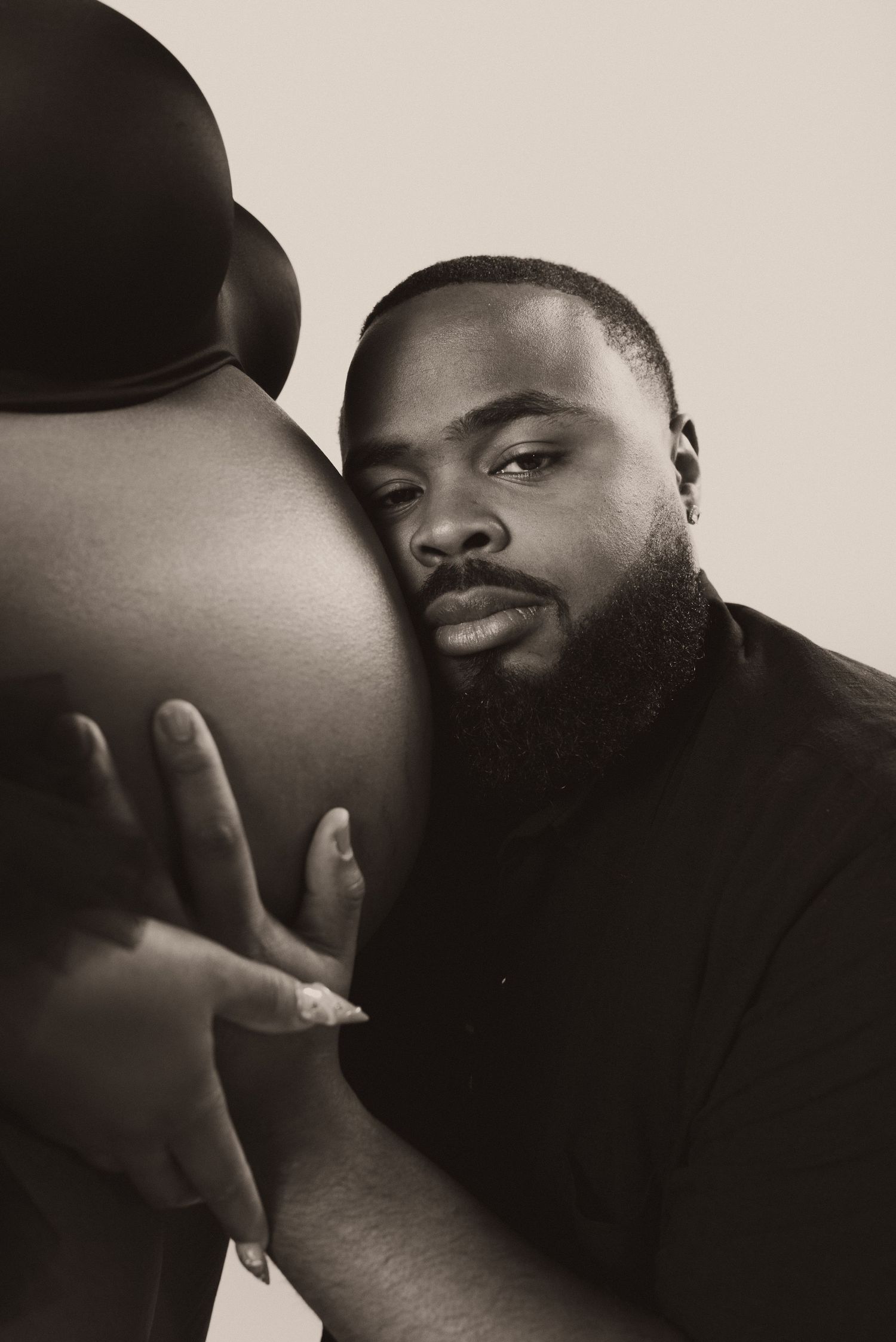 A bearded Black man tenderly rests his cheek against a pregnant woman's bare belly in a sepia-toned maternity portrait.
