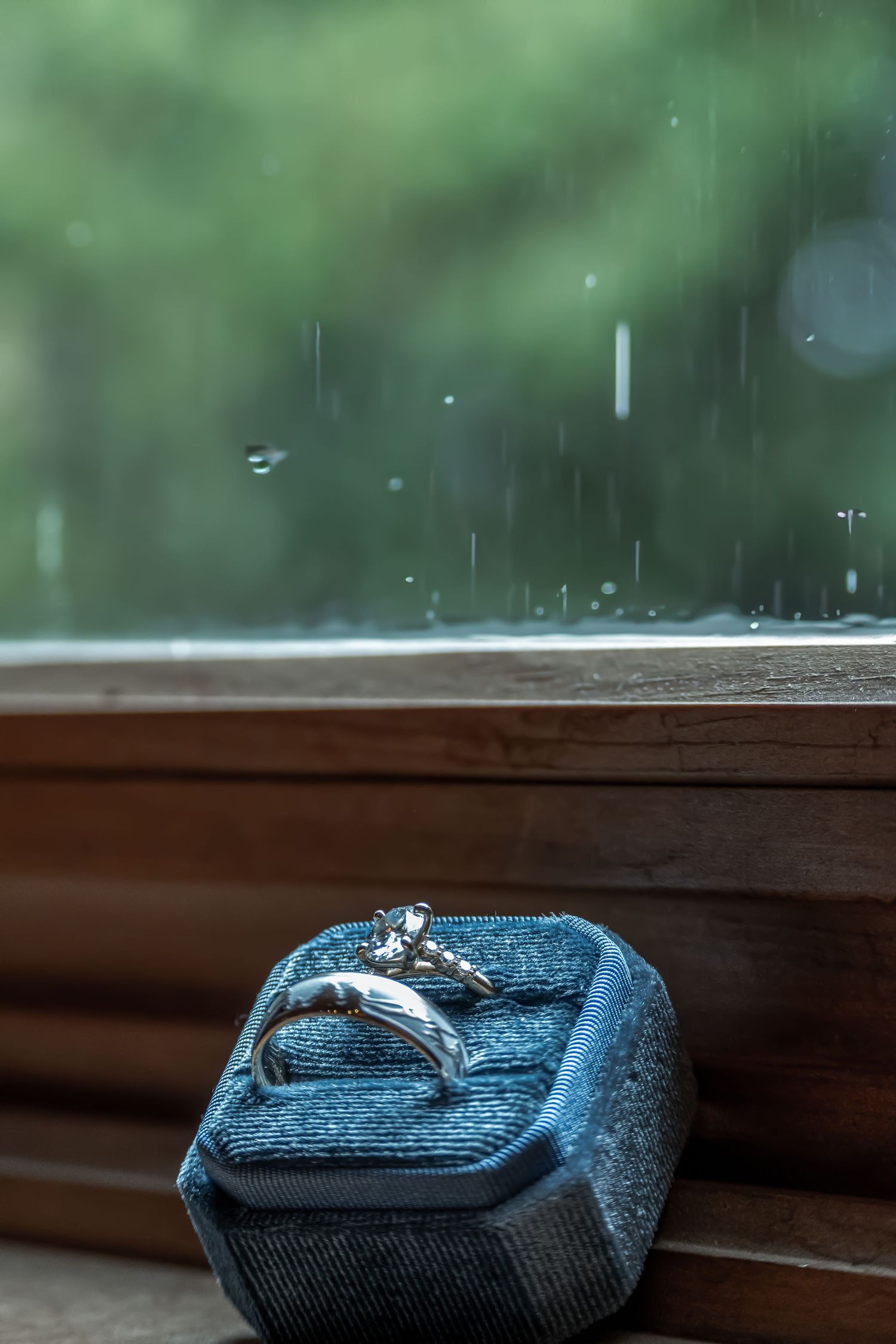 Diamond engagement ring on blue velvet ring box by a window on rainy day.