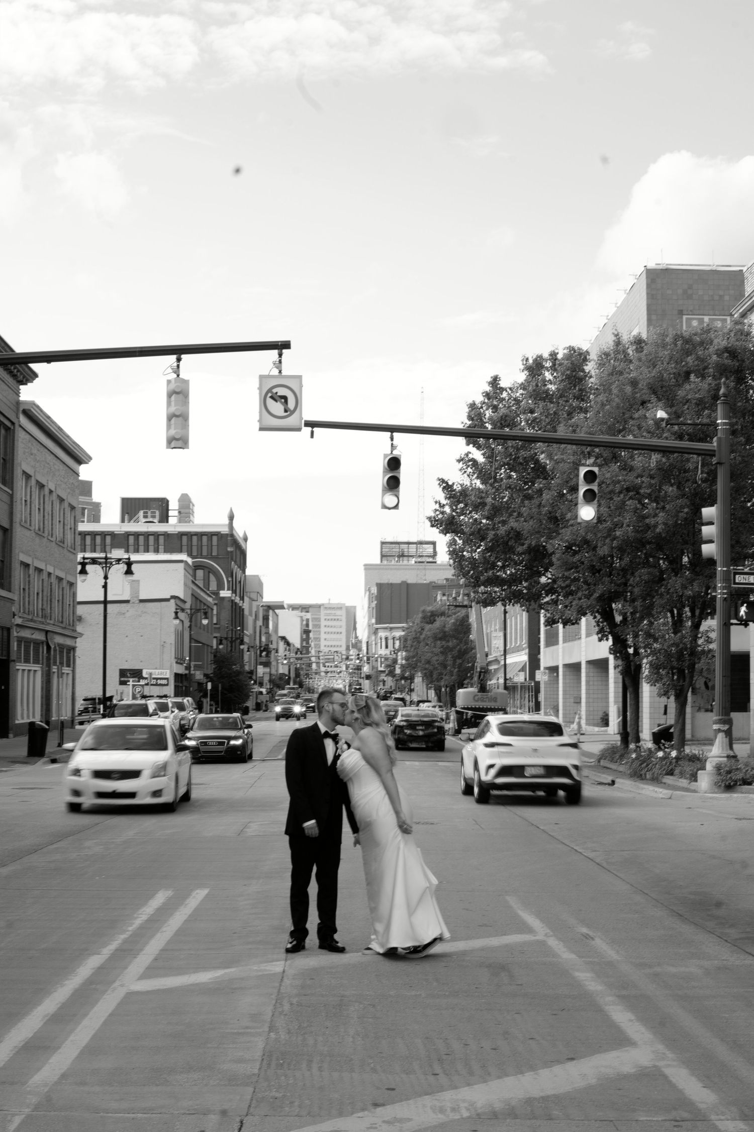 Black and white photo of a bride and groom kissing in the middle of a busy city street intersection.