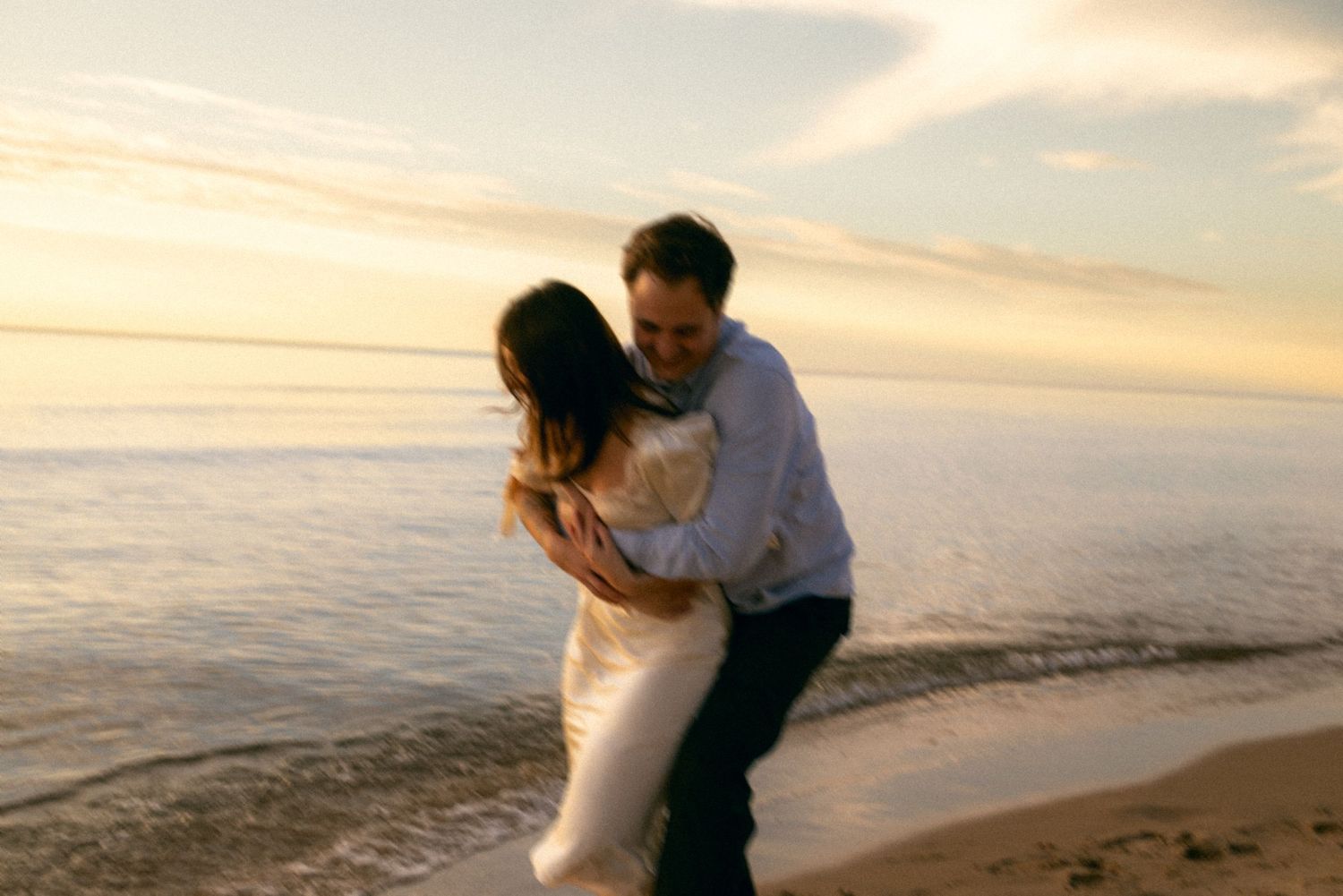 Romantic couple embracing on sandy beach during golden hour with ocean waves in background.