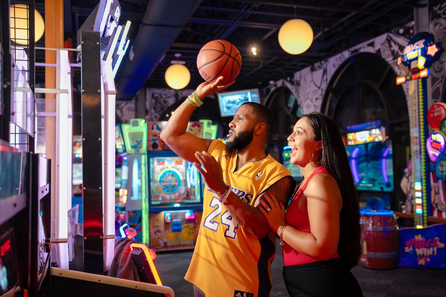 People enjoying arcade games with neon lights and basketball hoop in entertainment venue.
