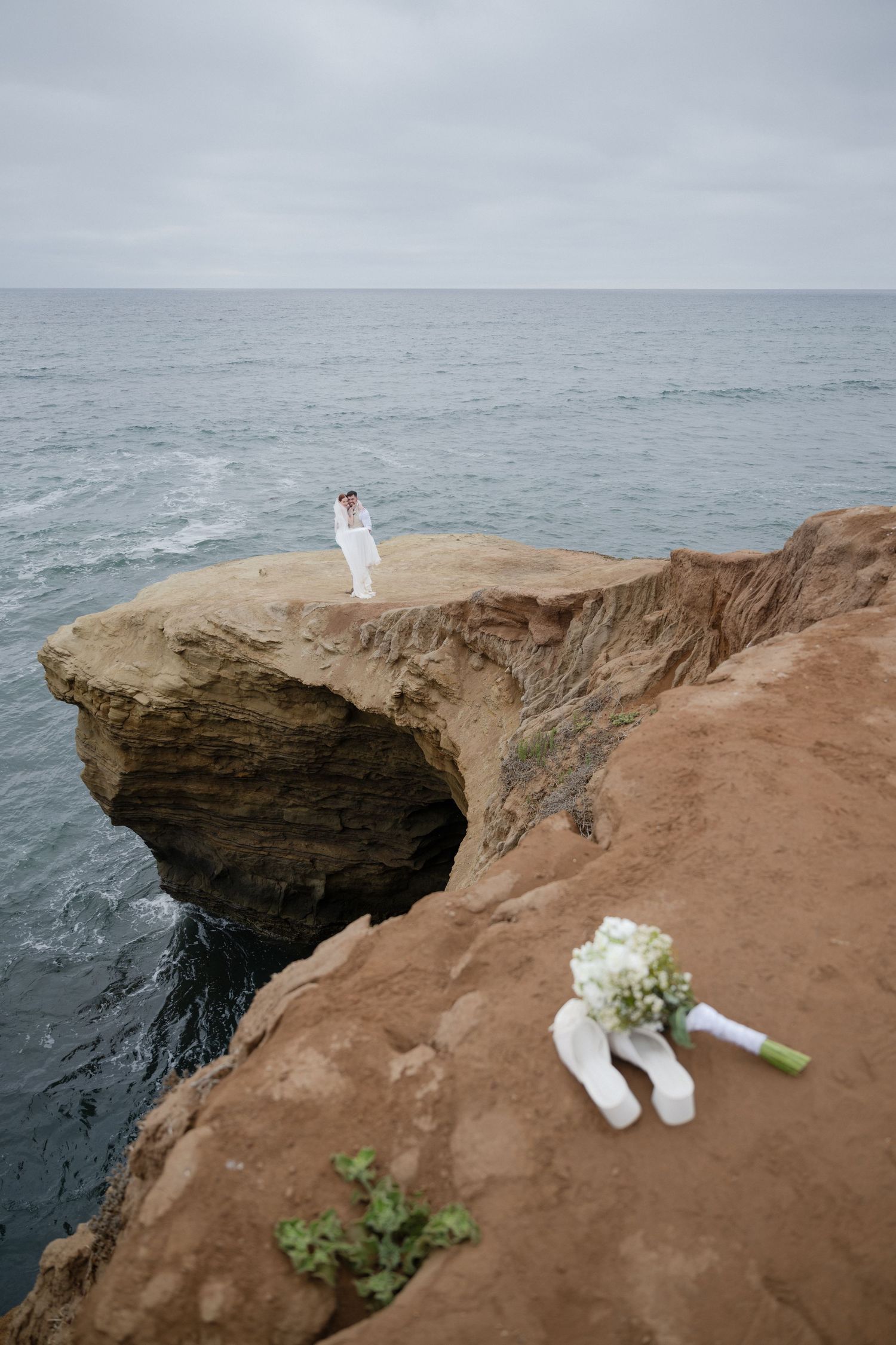 A bridal bouquet and shoes rest on rocky coastal cliffs overlooking the ocean.