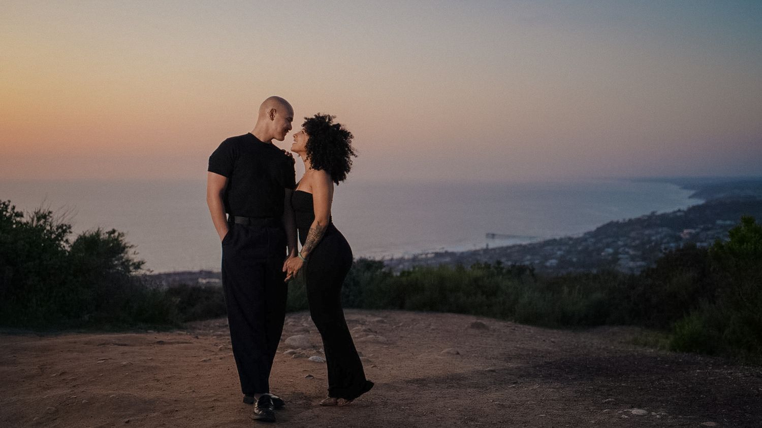 A couple stands in silhouette against a sunset sky overlooking coastal views.