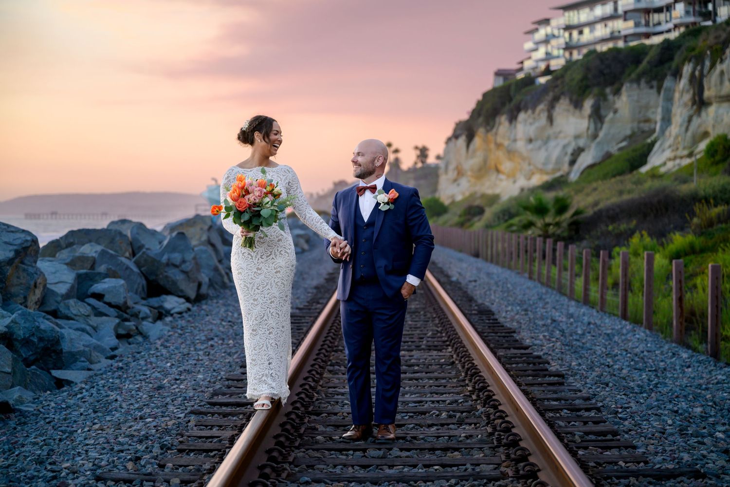 Couple walking along railroad tracks at sunset with cliffside homes in the background.