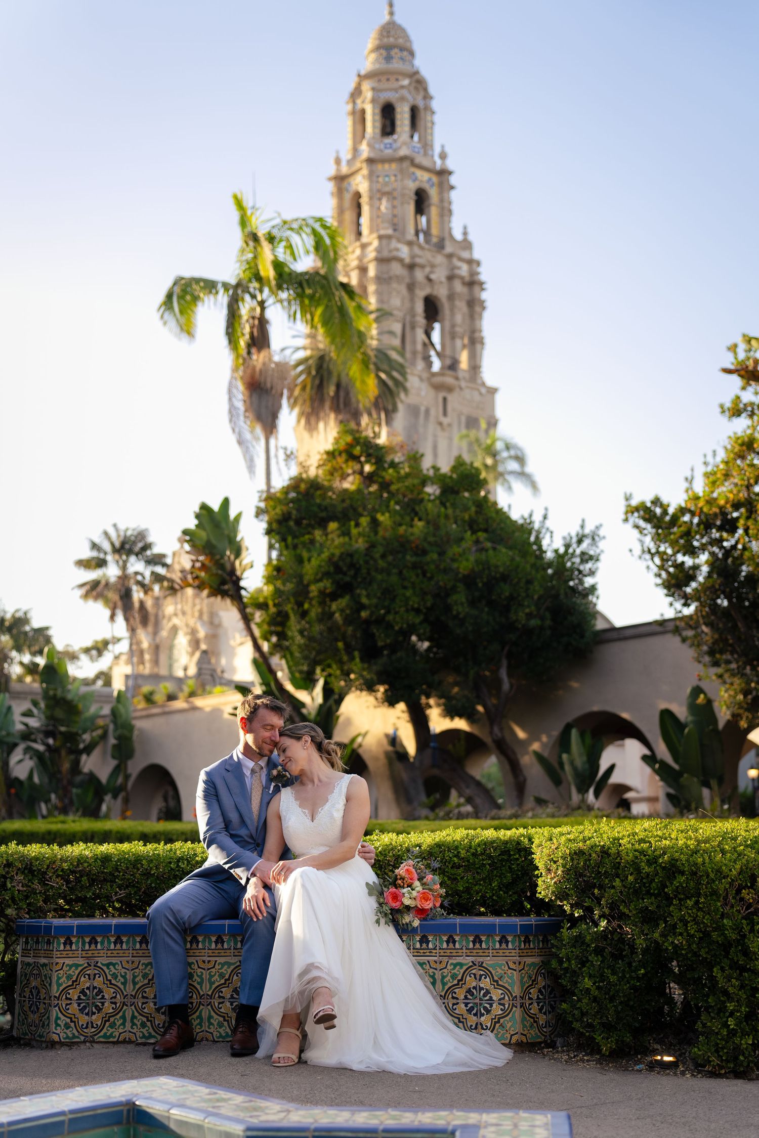 Romantic couple sitting by ornate fountain near Spanish colonial tower and palm trees.
