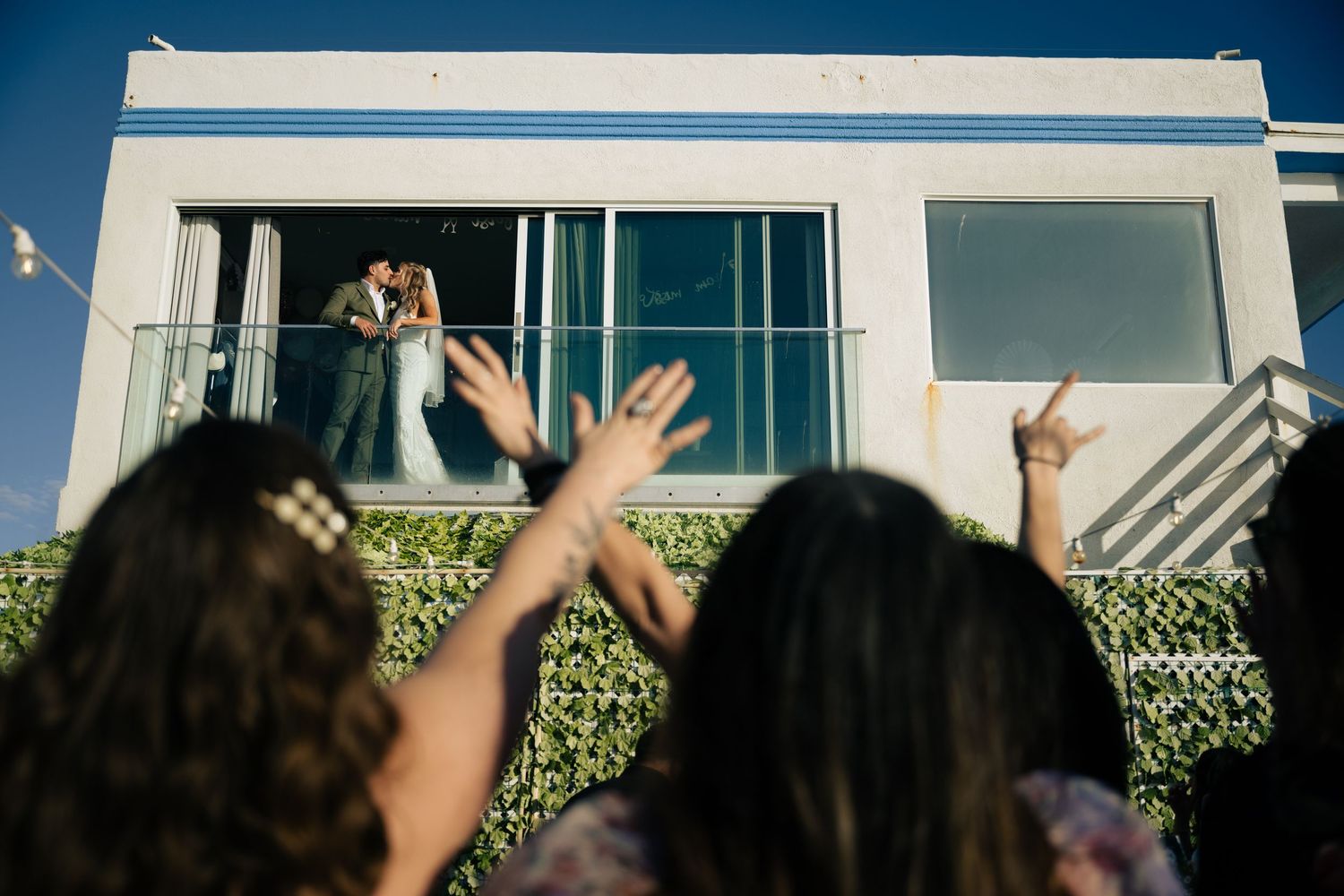 Wedding celebration with guests cheering from below as couple stands on white balcony.