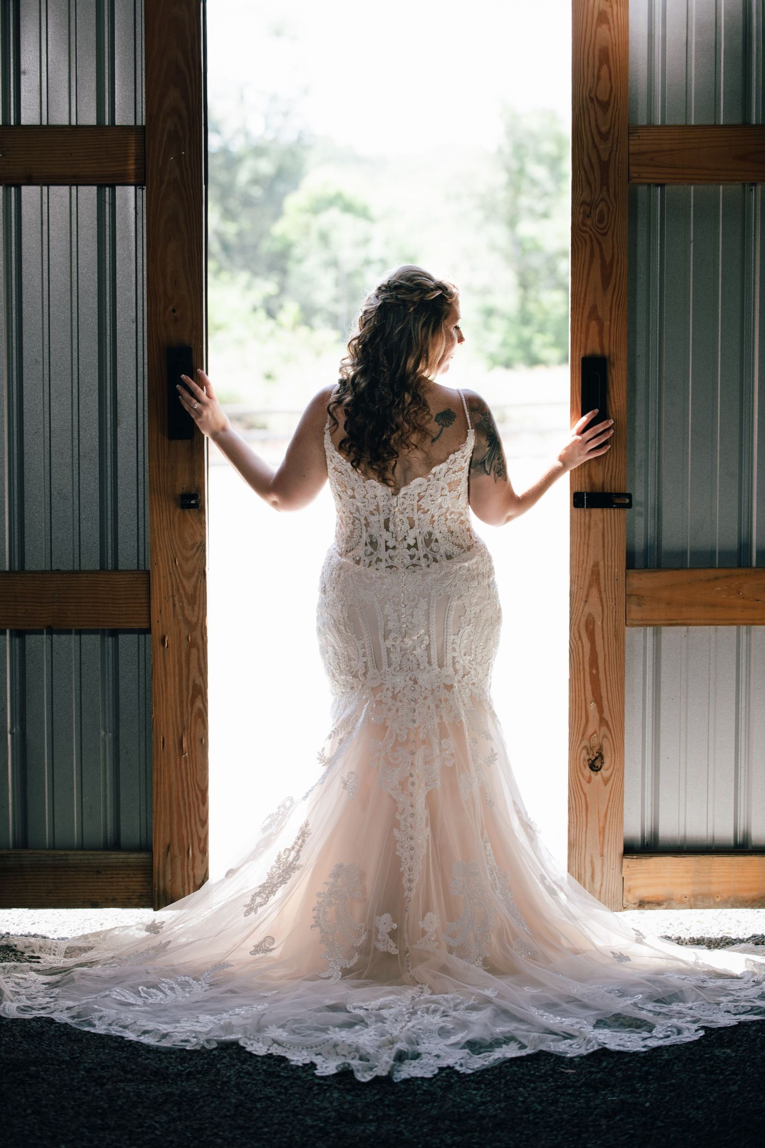 Bride in lace mermaid wedding gown stands backlit in open barn doors with flowing cathedral train.