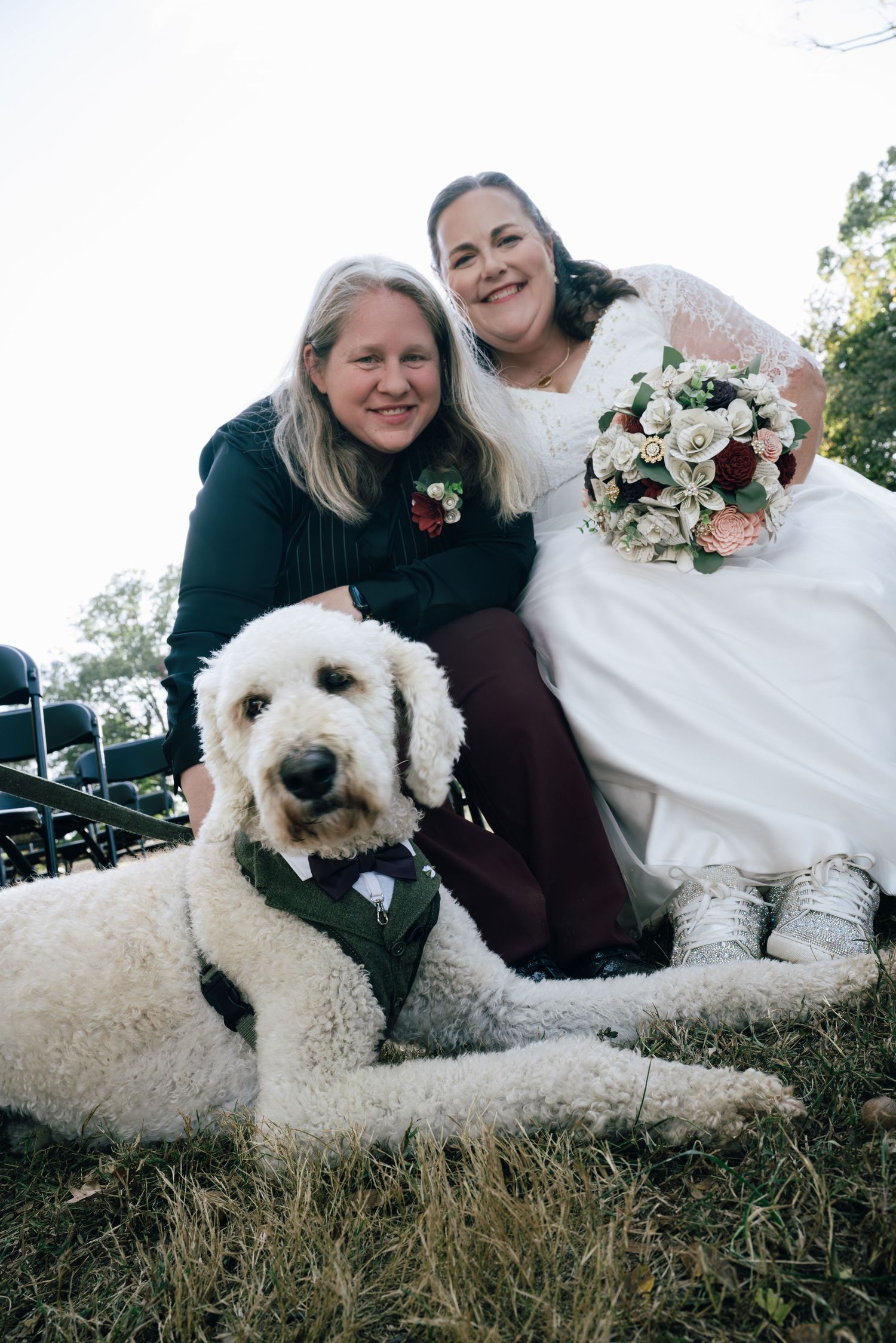 Two brides pose outdoors with large white fluffy dog wearing green vest at autumn wedding ceremony.