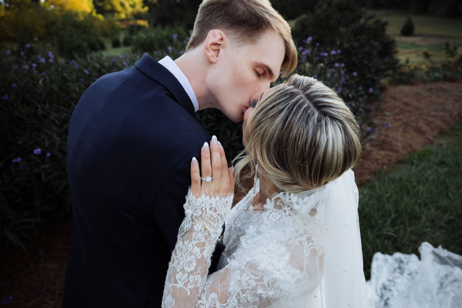 Groom in navy suit tenderly kisses bride in lace wedding dress on her forehead in garden setting.