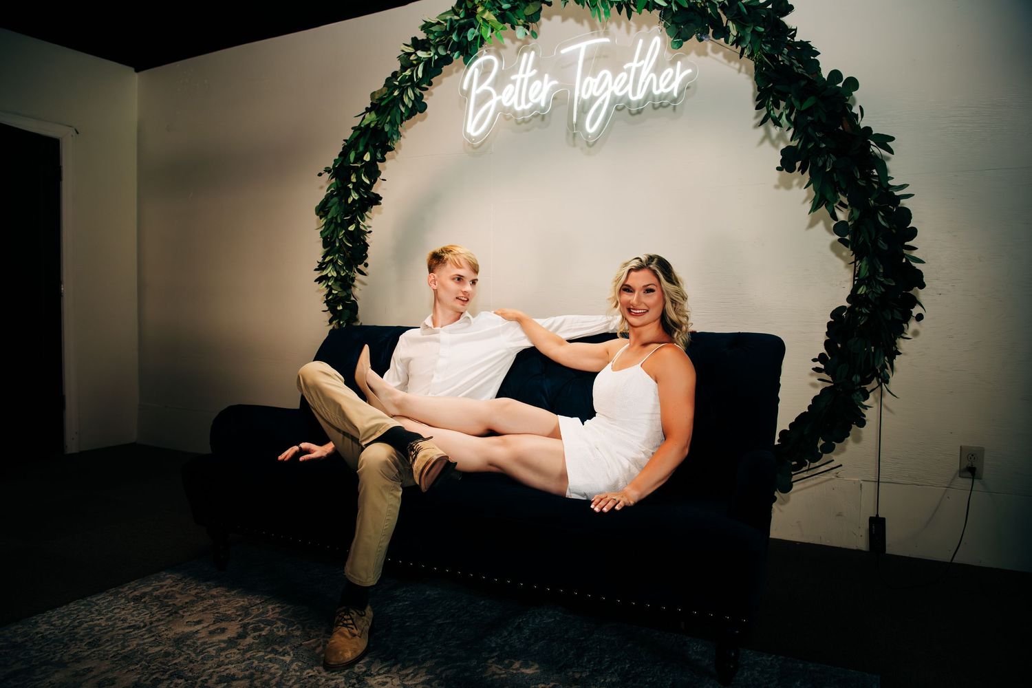 Couple poses on black velvet sofa beneath a Better Together neon sign with lush green floral wreath.