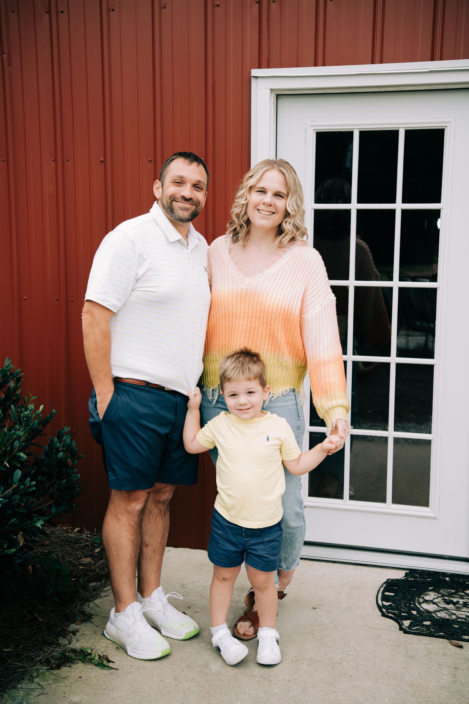 Family of three poses smiling outside red barn building, parents holding young boy's hands.
