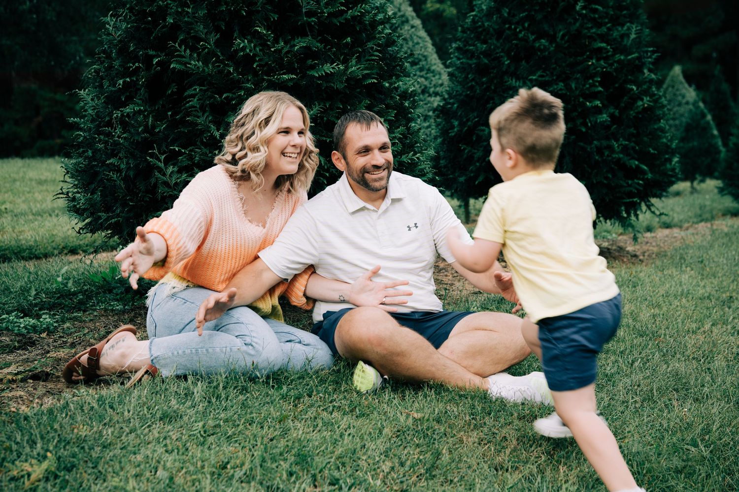 Happy family with young boy sits on green grass lawn surrounded by evergreen trees outdoors.