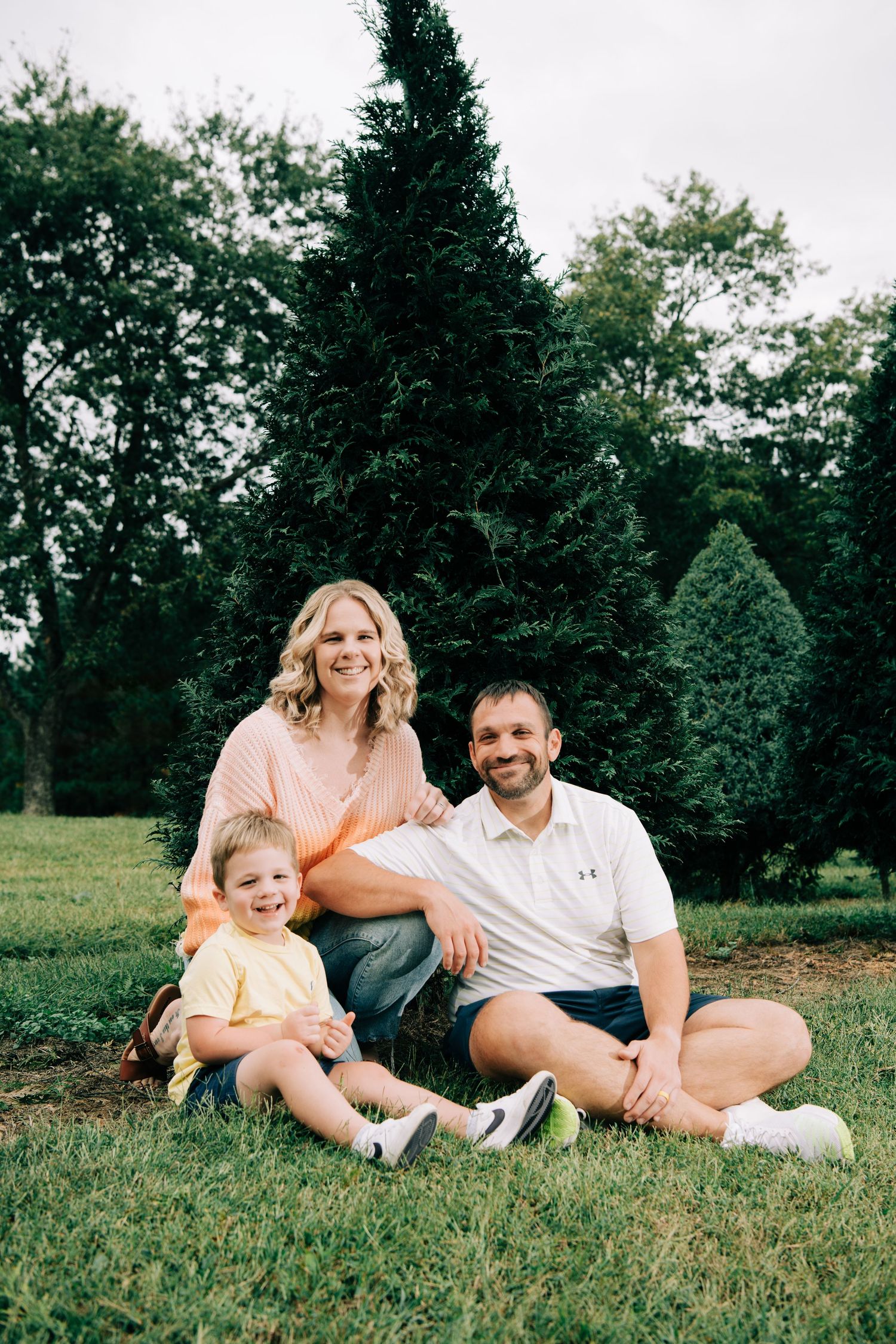 Happy family of three sits on green grass in front of tall evergreen trees.
