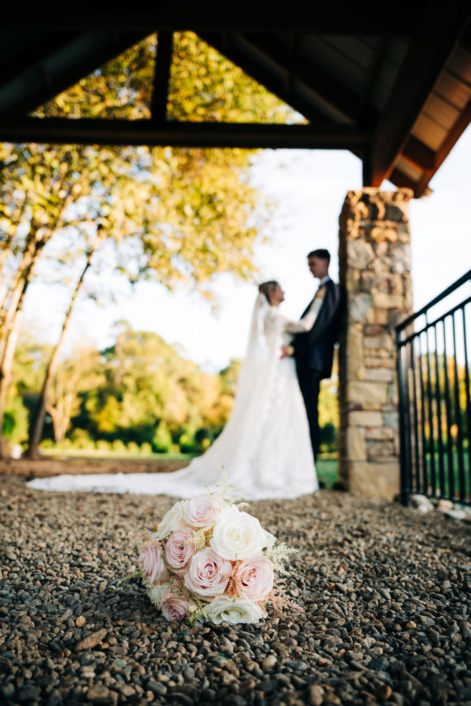 Bridal bouquet of pink and white roses in foreground with couple embracing under rustic stone pavilion.