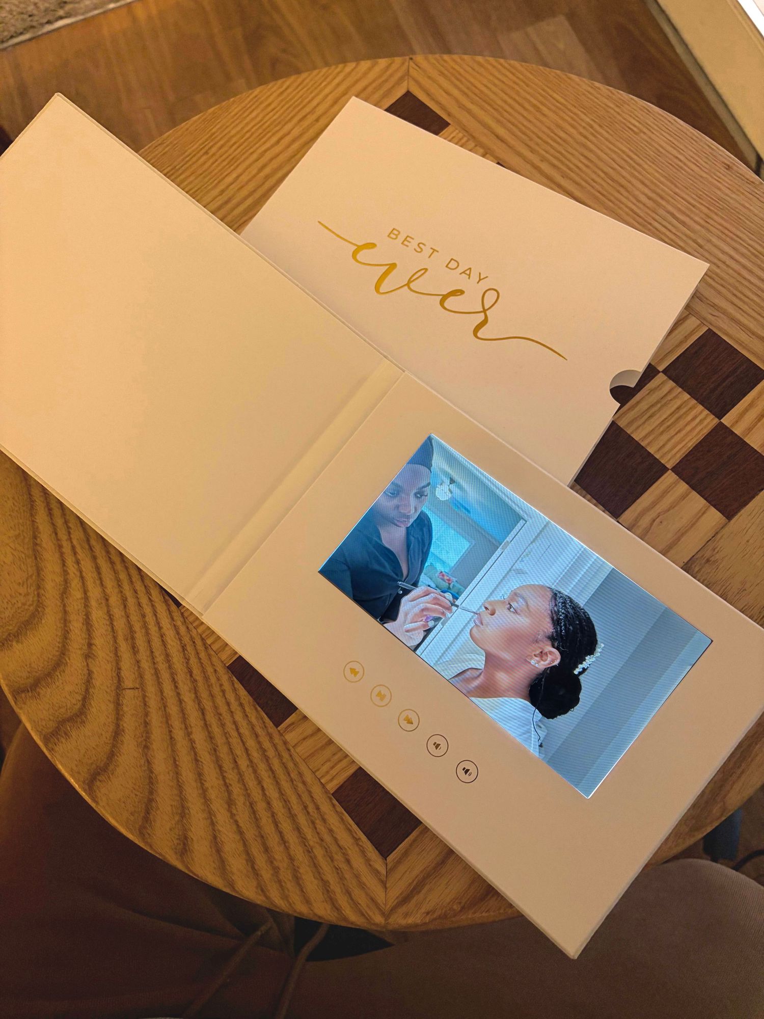 Elegant white video wedding book with gold Best Day Ever script displays couple video on wooden table.