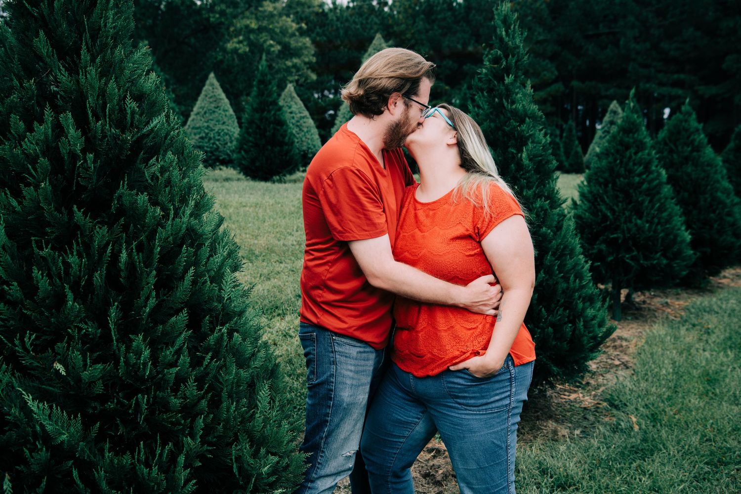 Couple in matching red shirts share a kiss among green pine trees outdoors.