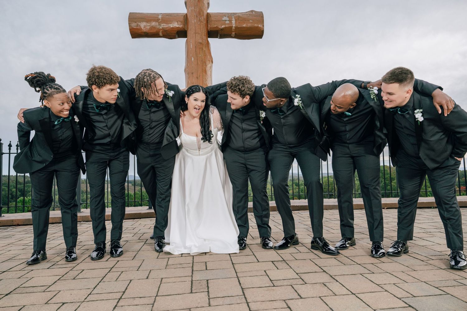 Bride in white gown poses with groomsmen in black suits before large wooden cross.