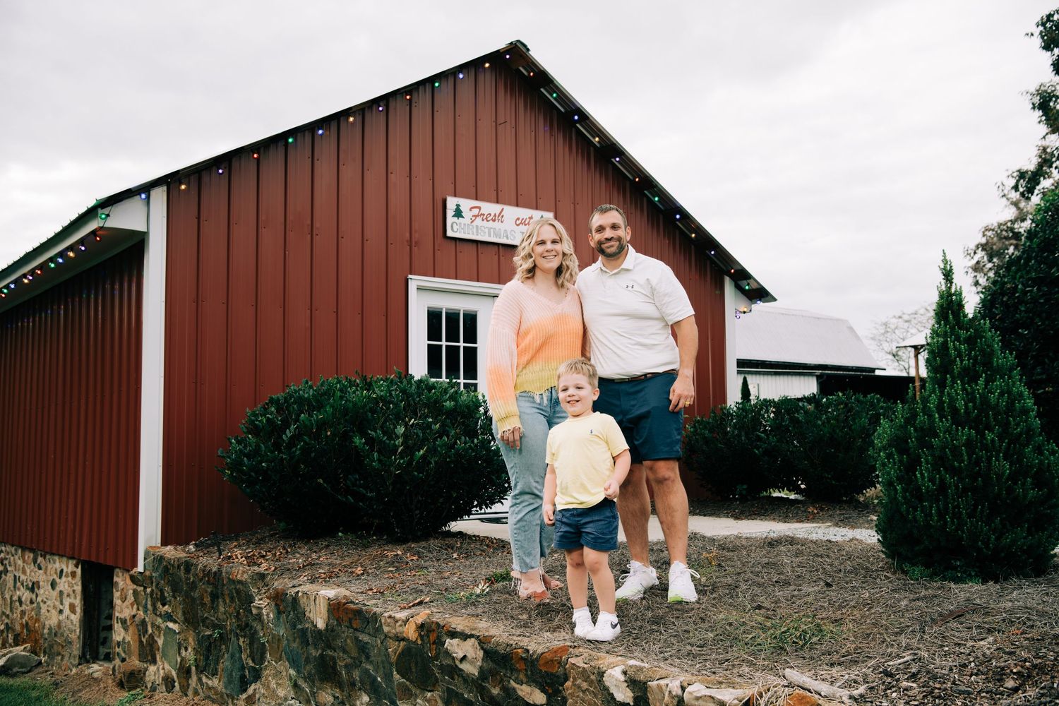 Family of three stands together smiling outside a red barn building on cloudy day.