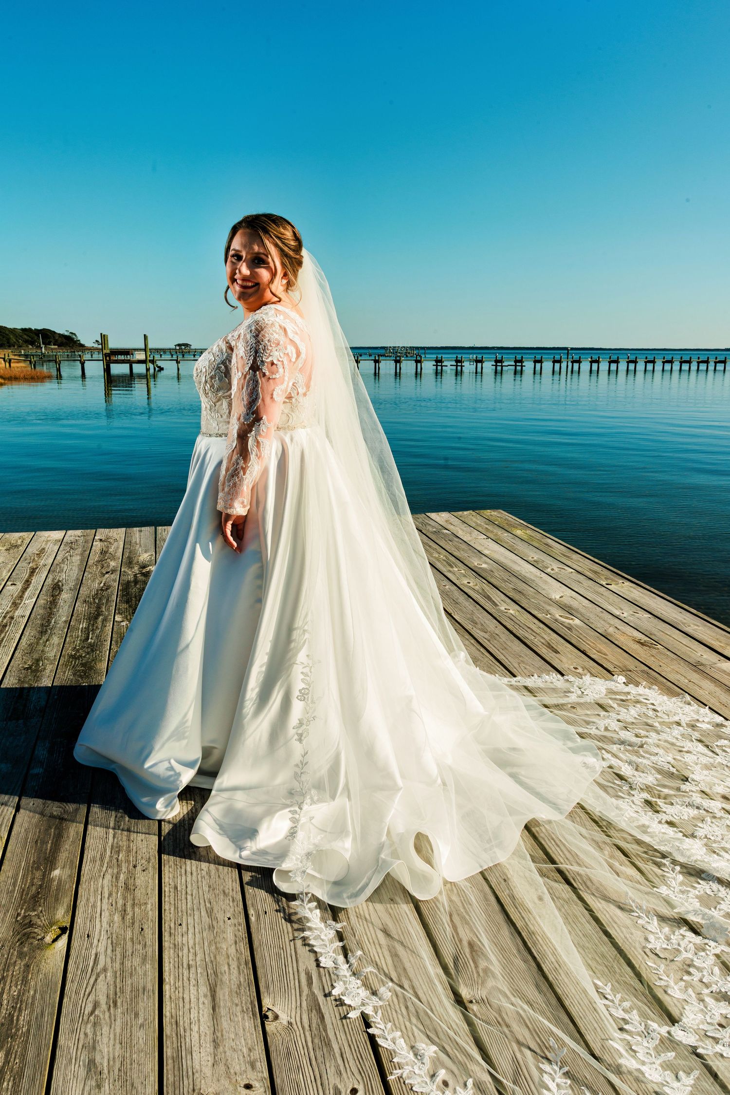 Bride in lace long-sleeve wedding dress with veil stands on wooden dock overlooking calm blue water.