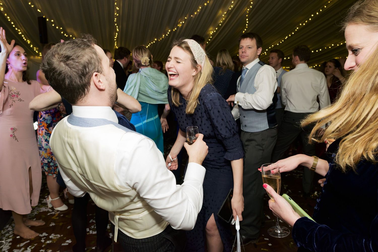 Wedding guests dance and celebrate on a crowded dance floor illuminated by warm string lights during an evening reception.