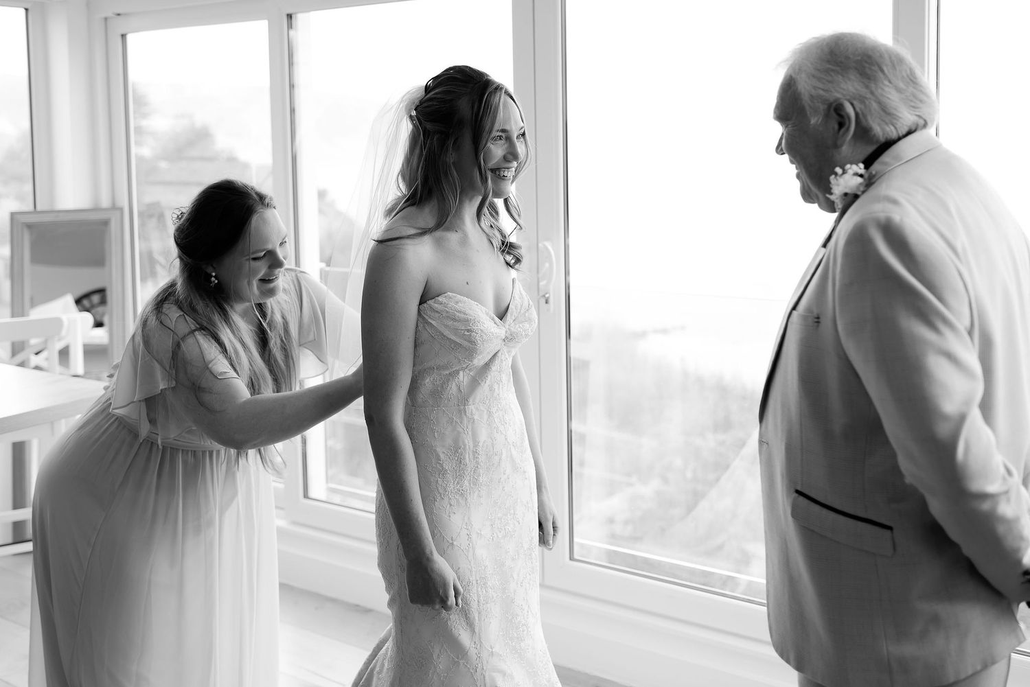 A bride being helped with her wedding dress while standing near a bright window in a black and white photograph.