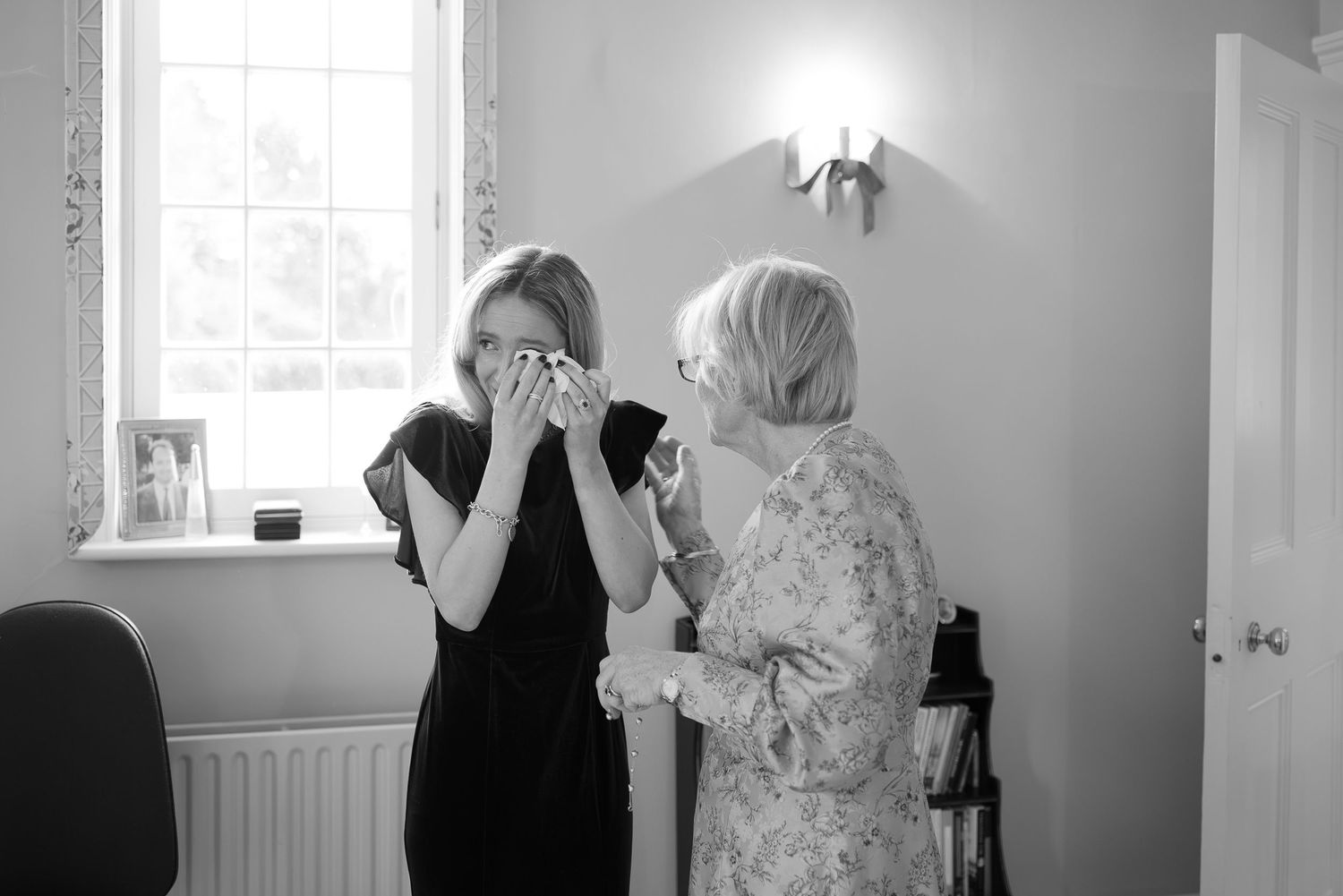 A bride in a wedding dress stands in a sunlit room during wedding day preparations.