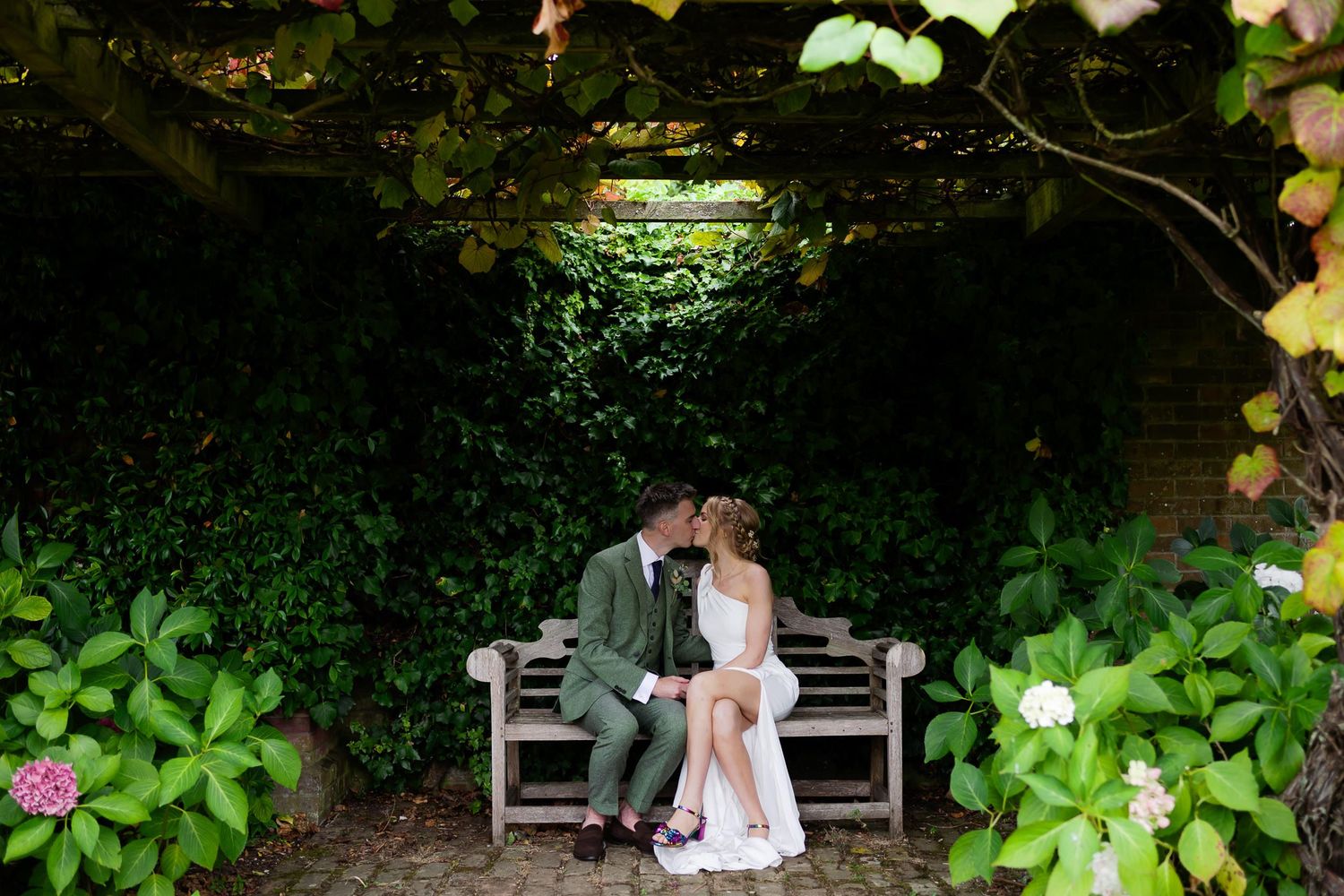 Newlyweds share intimate moment on garden bench surrounded by lush greenery.