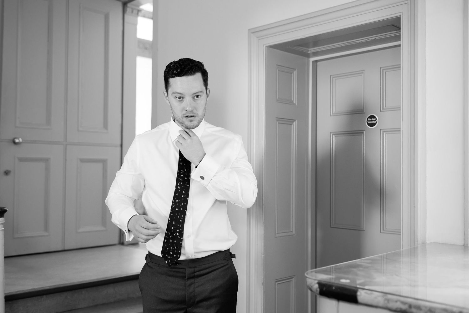 Man in formal attire adjusting his tie in a bright white room.