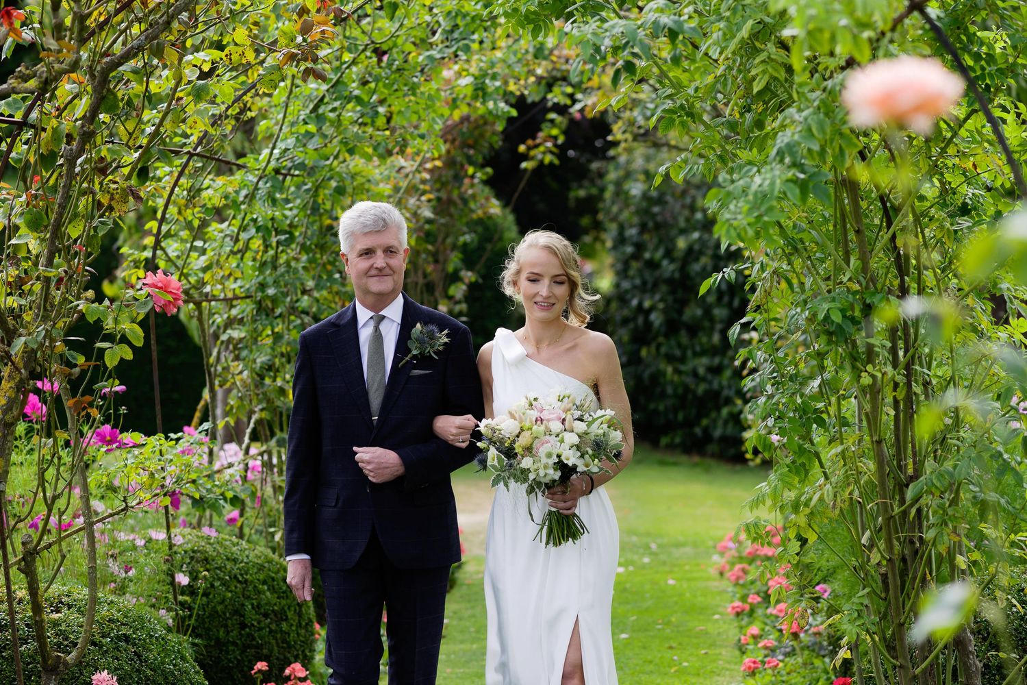 Classic father-daughter moment walking through garden pathway before wedding ceremony.