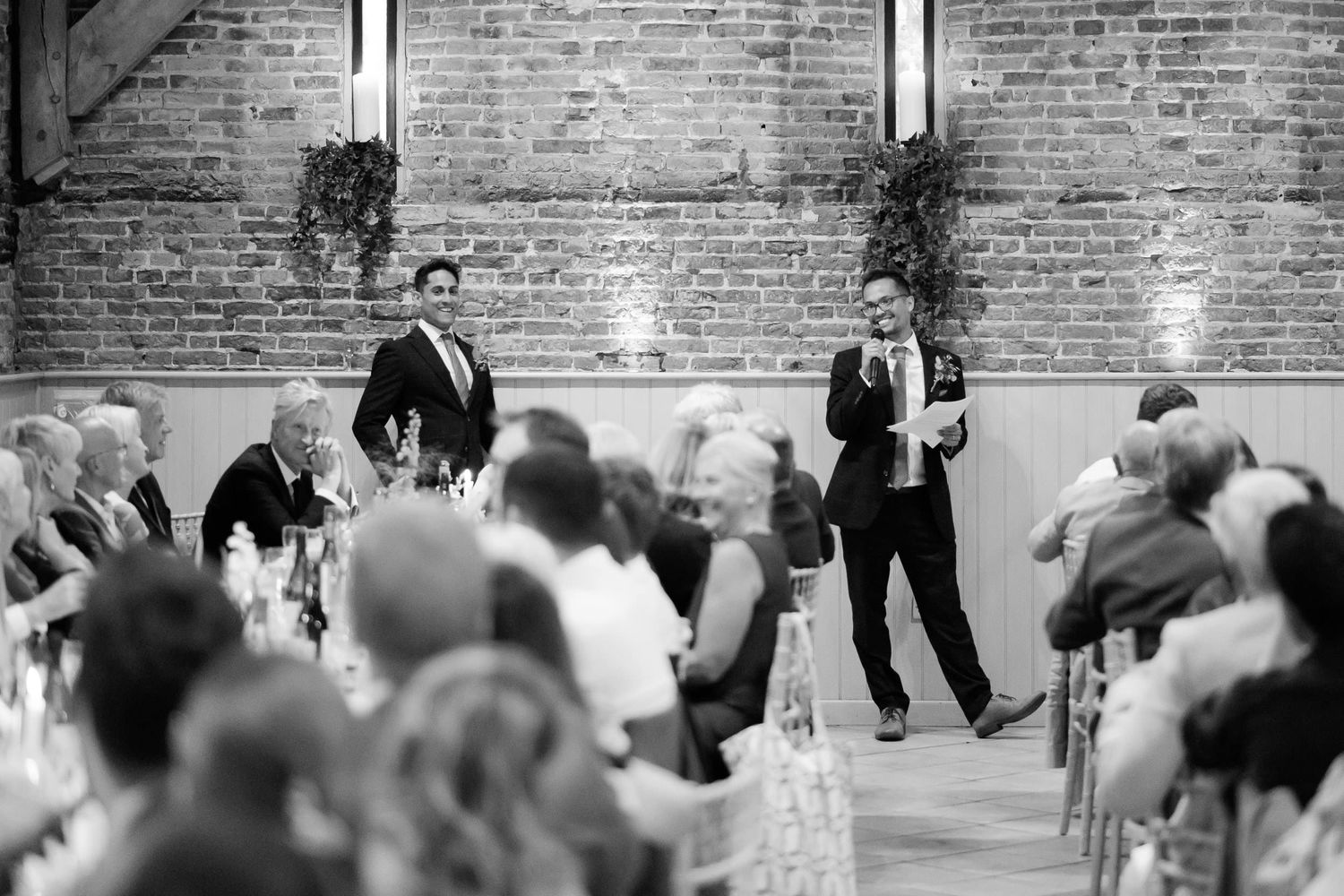 Wedding speeches captured in black and white with exposed brick backdrop and standing audience.