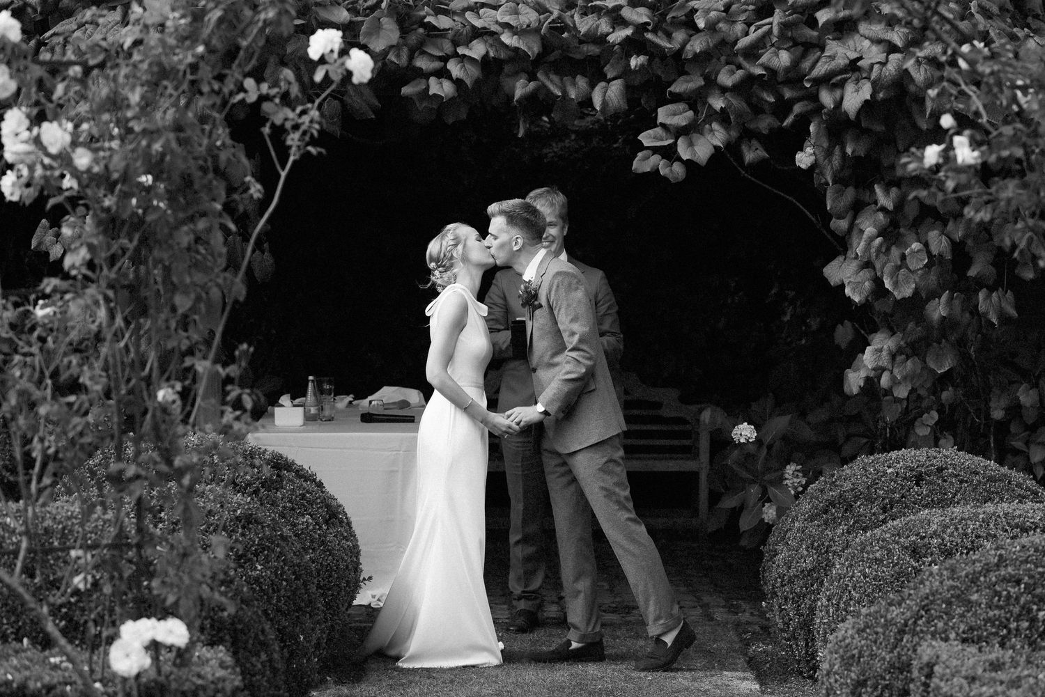 A couple shares an intimate moment in a garden setting with lush greenery and white flowers in the background.