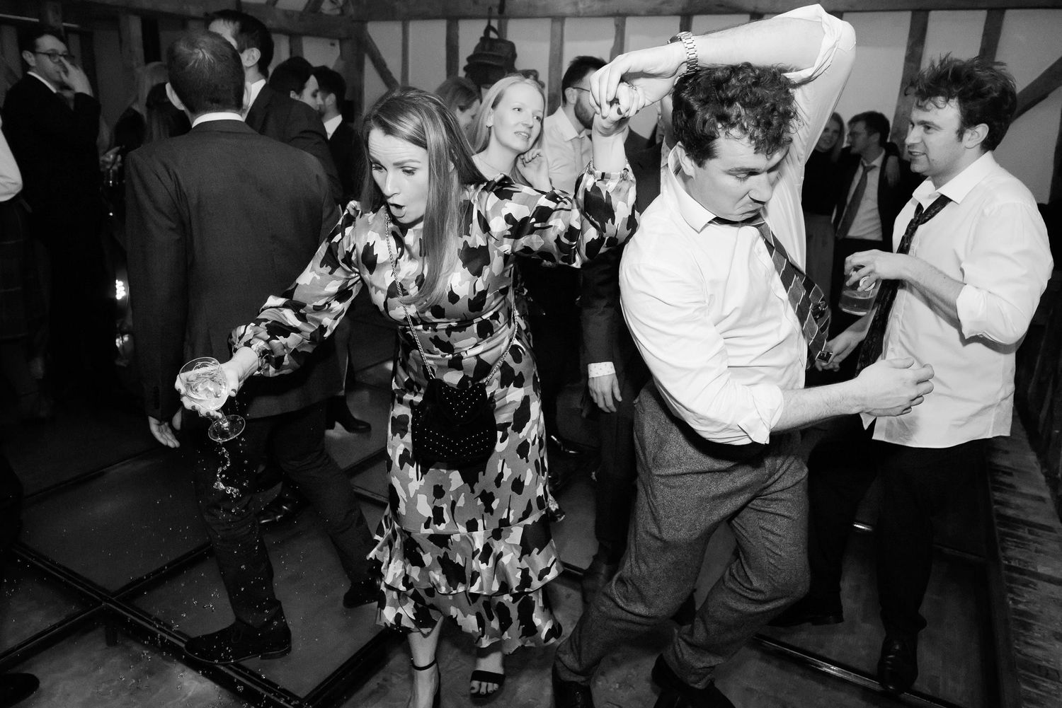 Wedding guests dancing enthusiastically at a reception with people wearing floral dresses and formal attire.